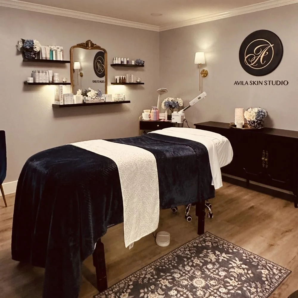 A treatment room at Avila Skin Studio with a massage table covered in black and white linens, surrounded by shelves with skincare products, a mirror, flowers, and soothing lighting.