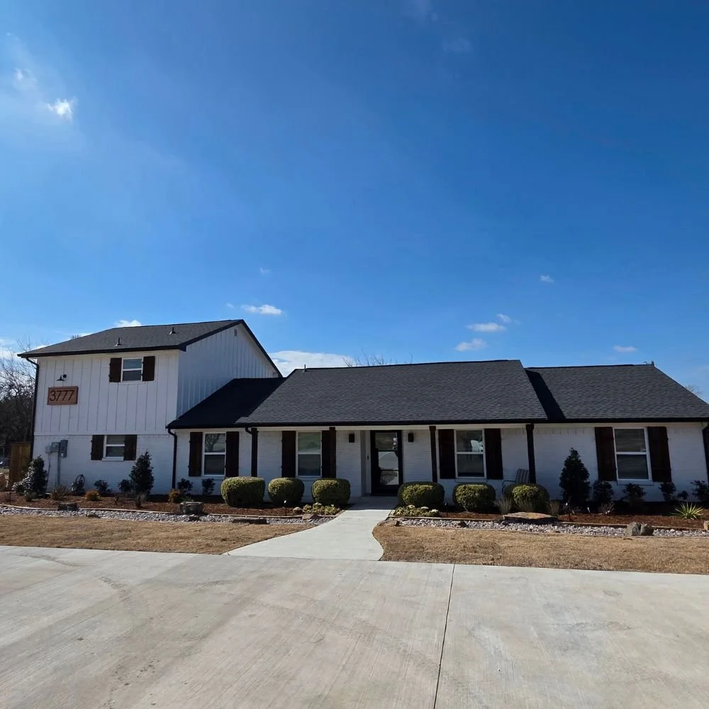 Avila Skin Studio is inside this two-story white house with black shutters, a dark gray roof, a small front porch, surrounded by neatly trimmed bushes, with a sidewalk leading to the front door, under a bright blue sky.