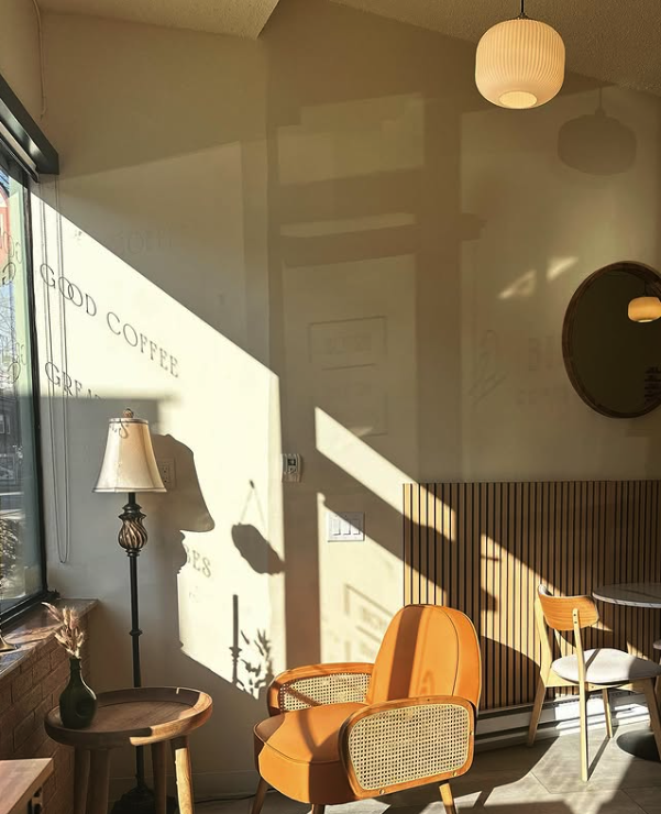 Sunlit interior of a cozy coffee shop with a warm color palette, featuring a mustard chair, a small wooden table with a vase, a floor lamp, and a round wall mirror.