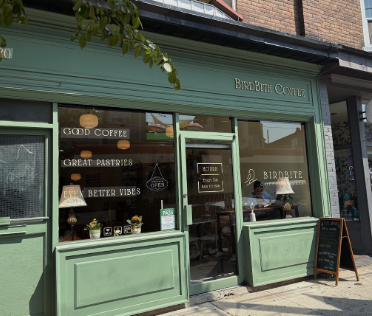 Green storefront of Birdbite Coffee with large window and door, chalkboard sign outside, displaying coffee shop interior with tables, chairs, and hanging lamps.