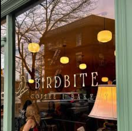 View of a bakery named Birdbite from outside the window with hanging ceiling lamps, a woman with curly hair, and a table lamp inside.