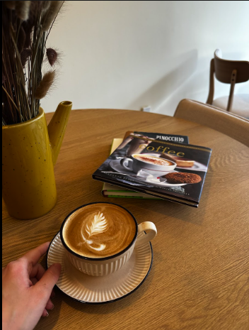 Cup of coffee with latte art on a wooden table, with a hand holding the cup, and a vase with dried flowers nearby. In the background, there are magazines and a chair.