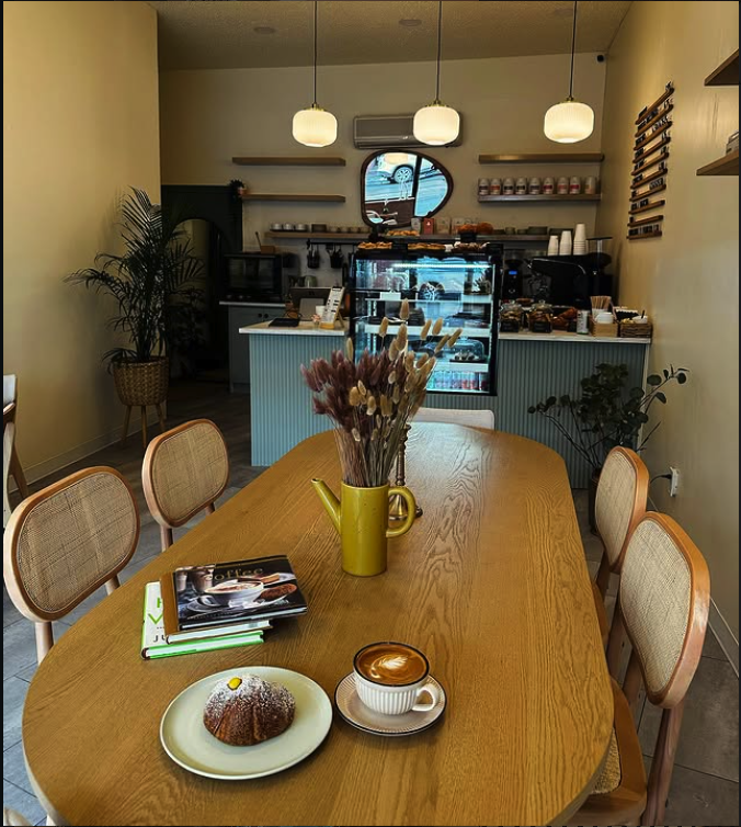 Coffee shop interior with a wooden table holding a book, a spoon, a pastry, and a cup of cappuccino, with a decor vase of dried flowers, and a display counter in the background.