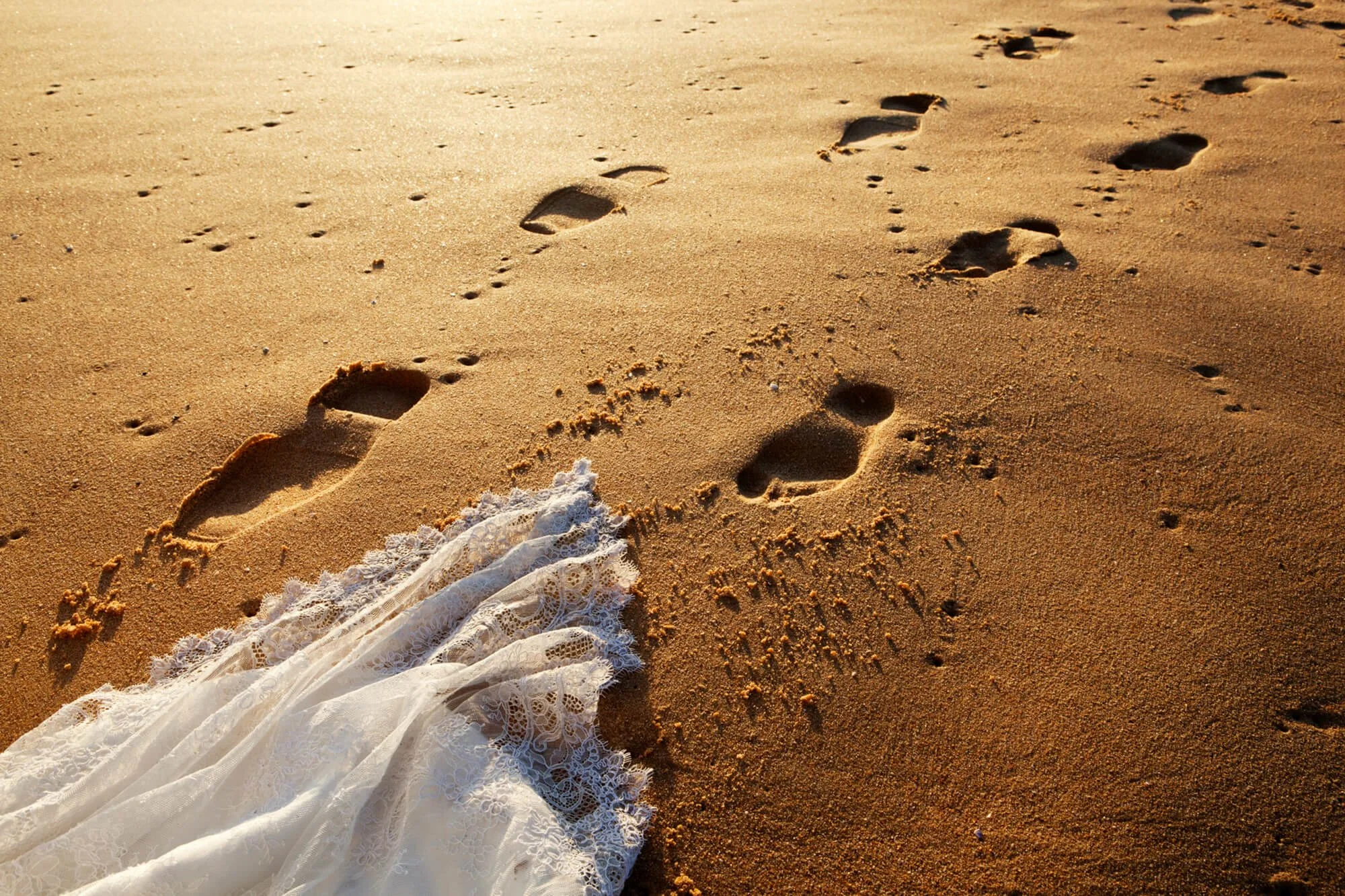 Trail of bare footprints in golden sand leading up to the bride's lace wedding dress train, soft evening light across Putty beach, Killcare