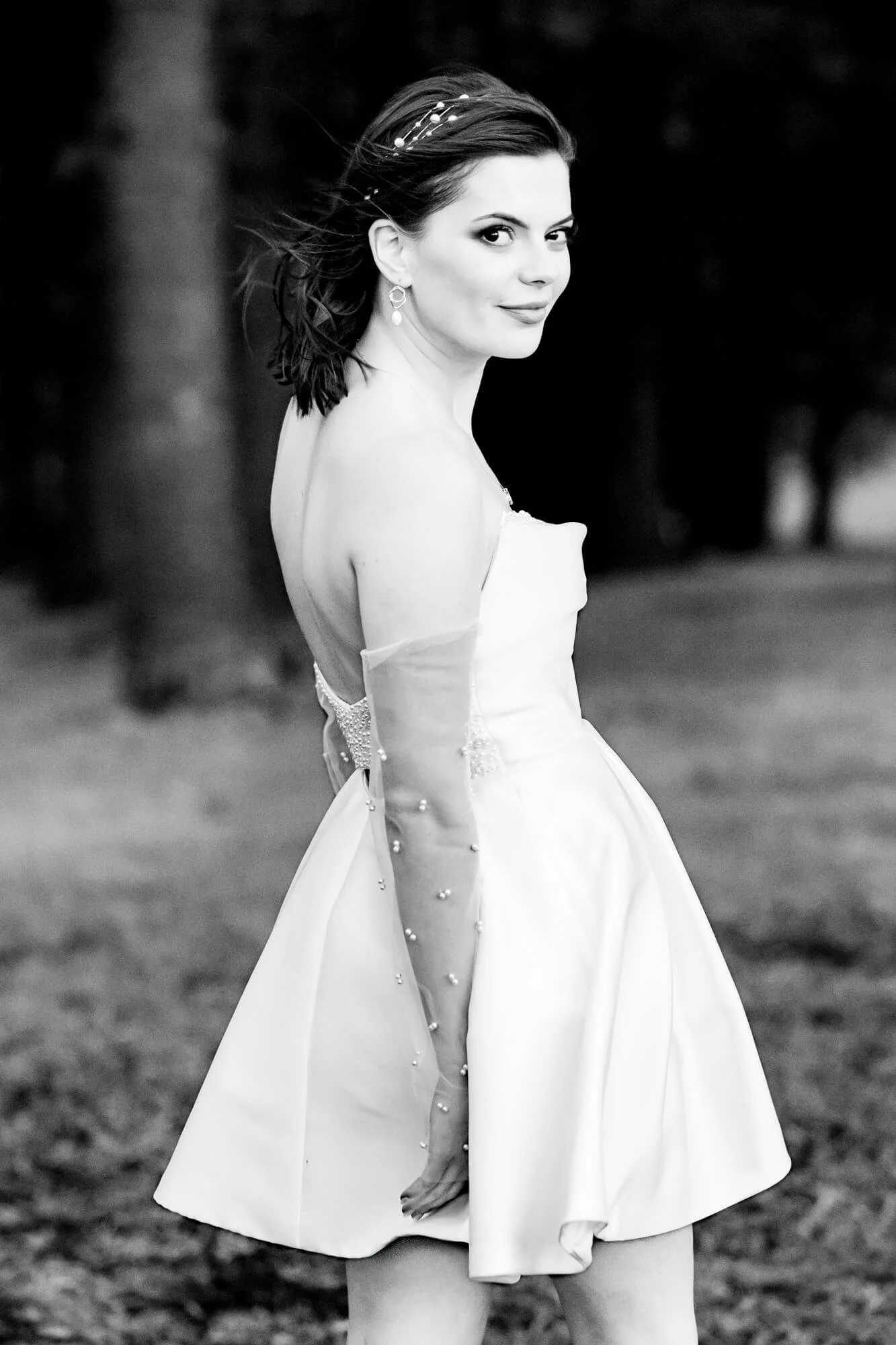 Bride glancing back with a soft, candid smile, her short tulle-and-pearl wedding dress and windswept hair set against a row of trees, King Edward Park, Newcastle.