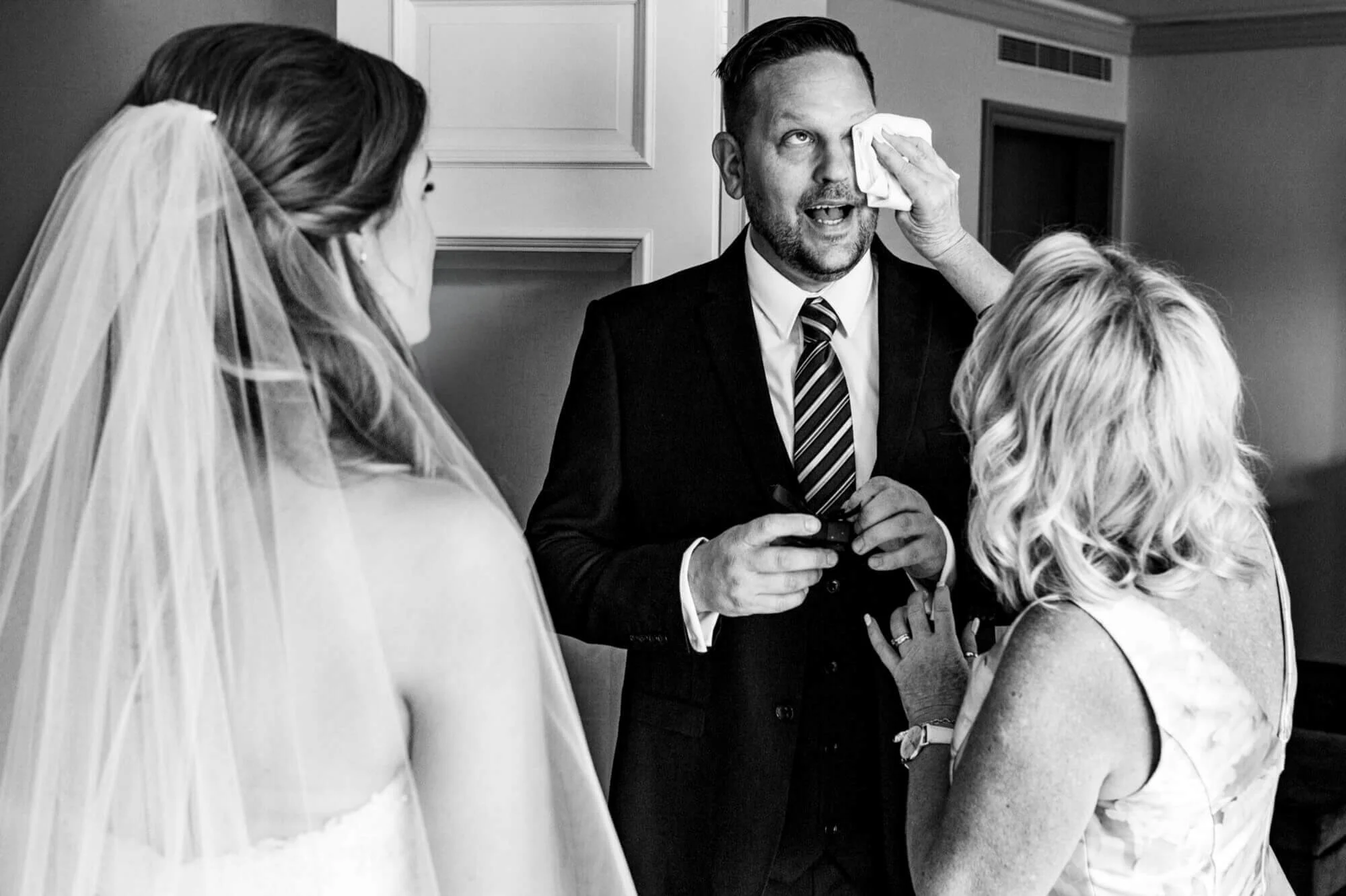 Bride's father wiping away tears of emotion as he sees her in her wedding dress for the first time, captured in black and white.
