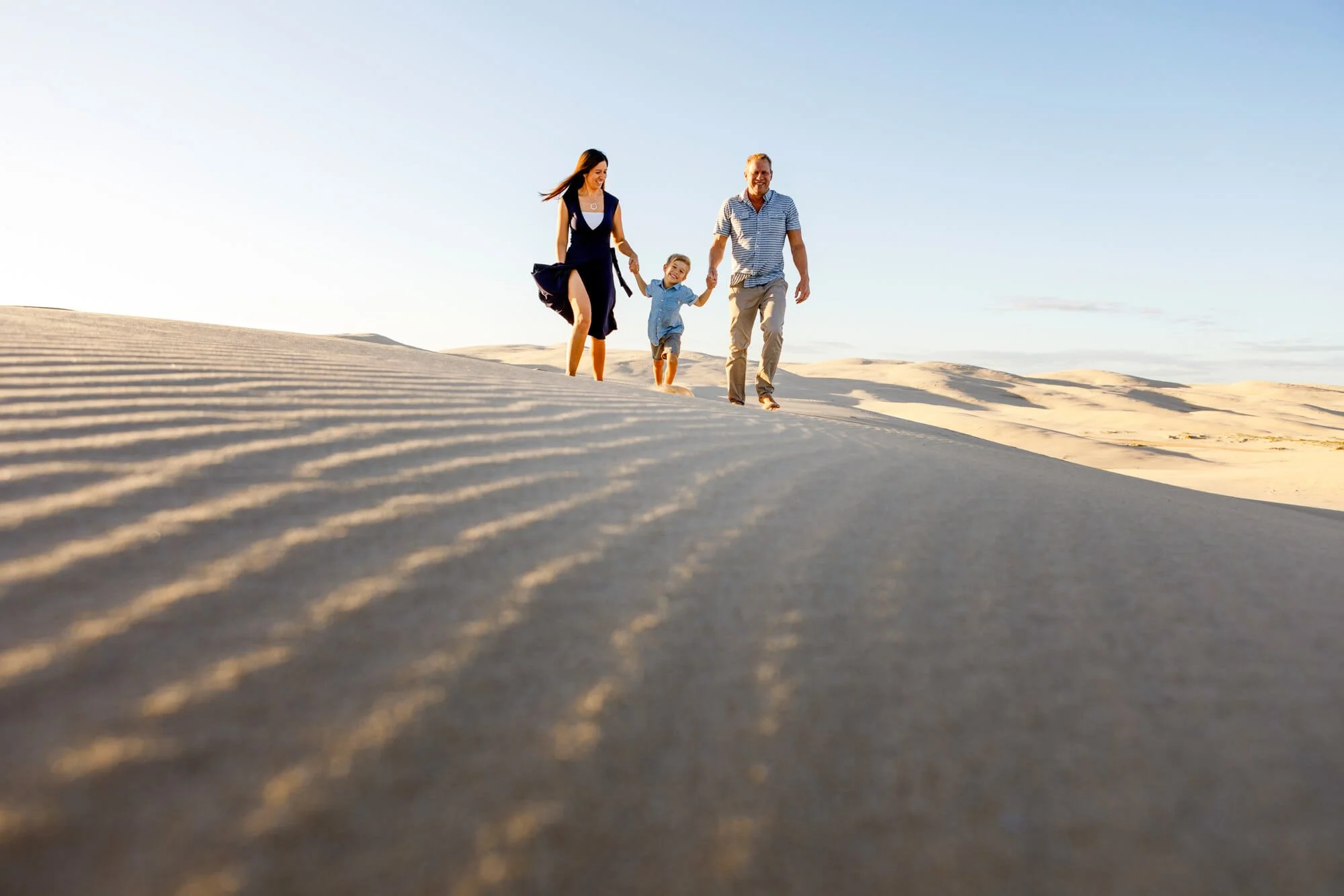 Parents walking hand in hand with their young child along a rippled sand dune at sunset, Anna Bay sand dunes, Port Stephens.