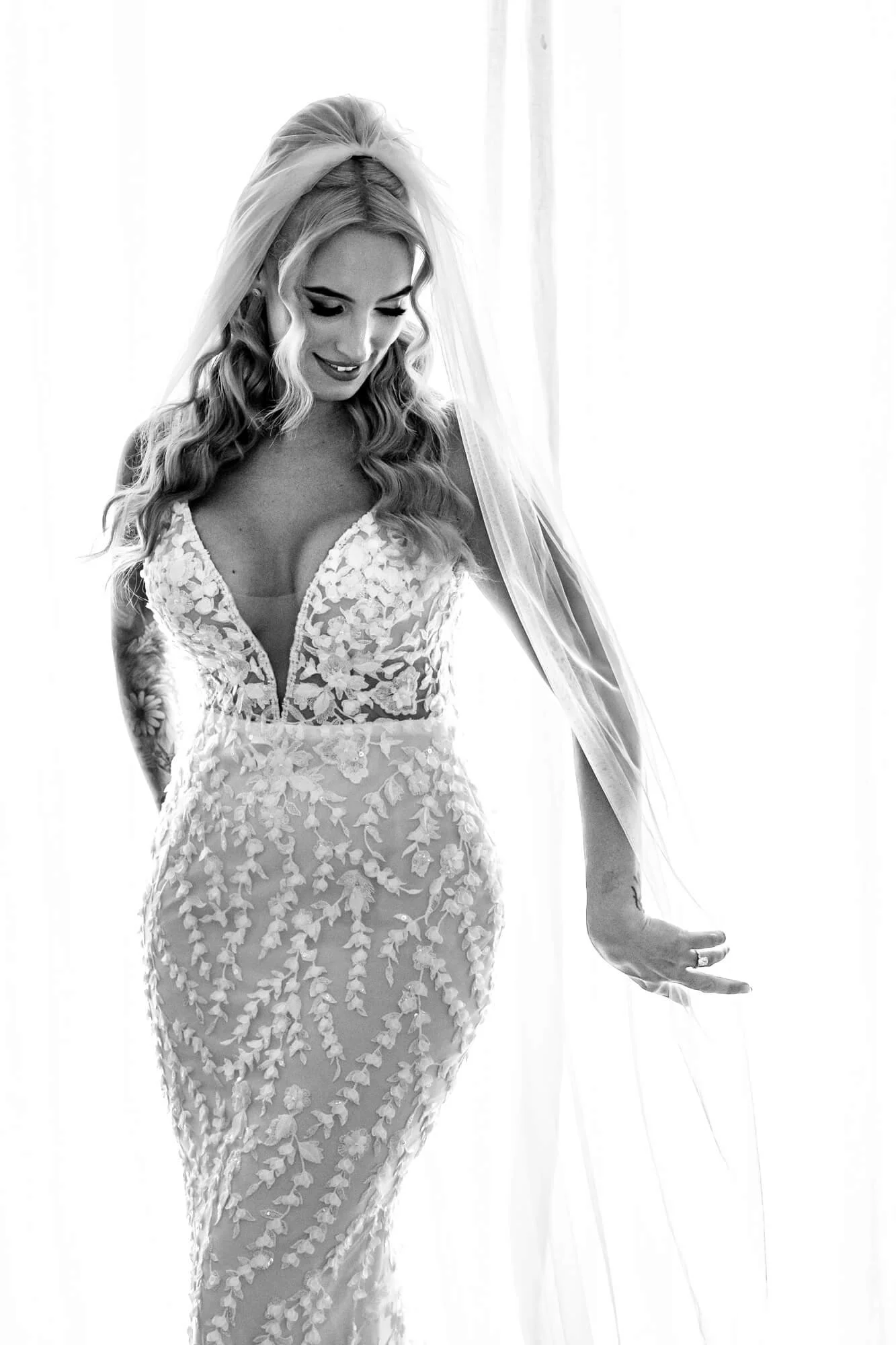 Black and white portrait of a bride in a fitted lace gown with a long cathedral veil, smiling softly as she stands by a window in natural light.