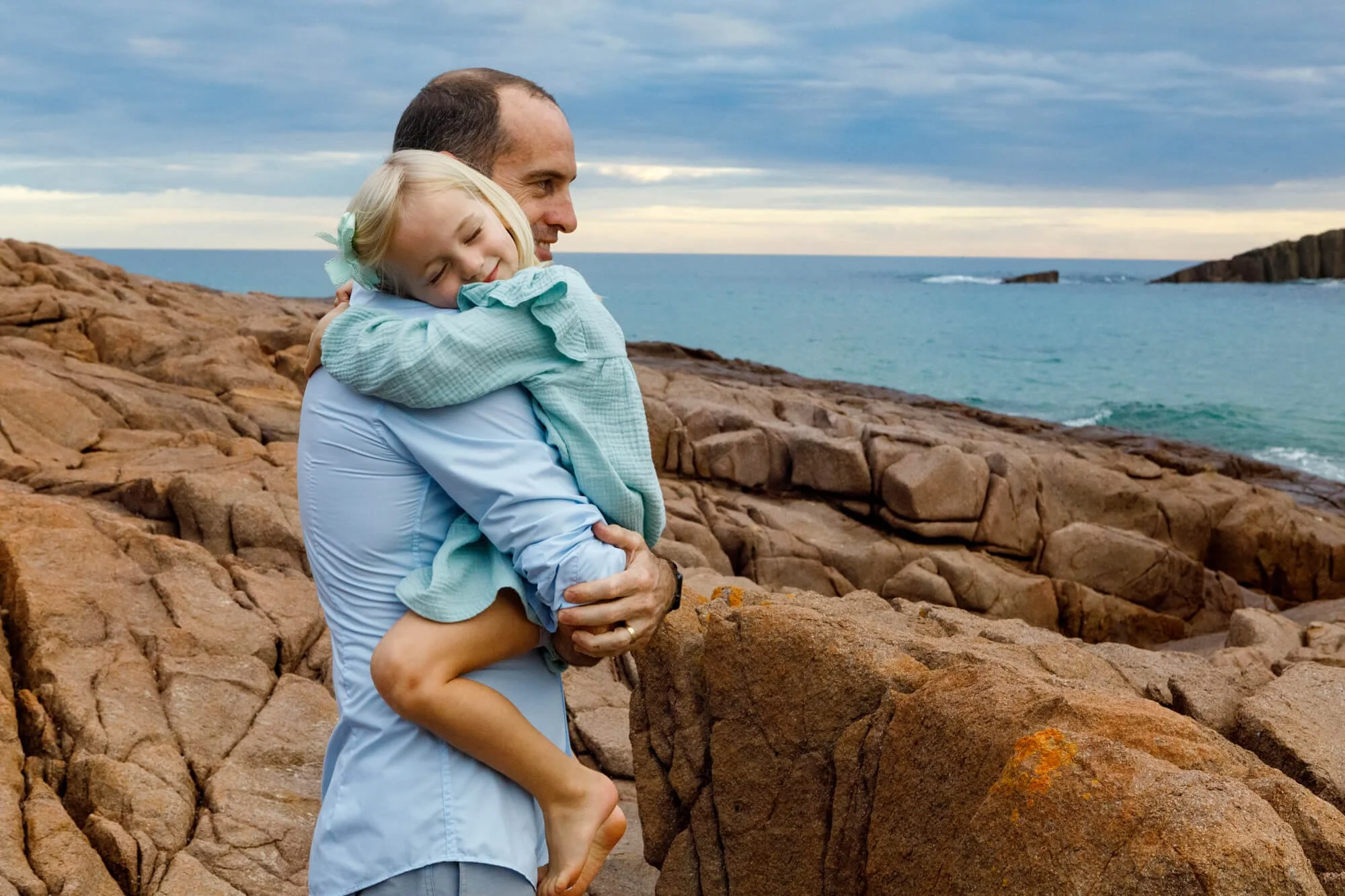 Dad carrying his young daughter in a tight hug on coastal rocks above turquoise water at sunset, Kingsley Beach, Boat Harbour.