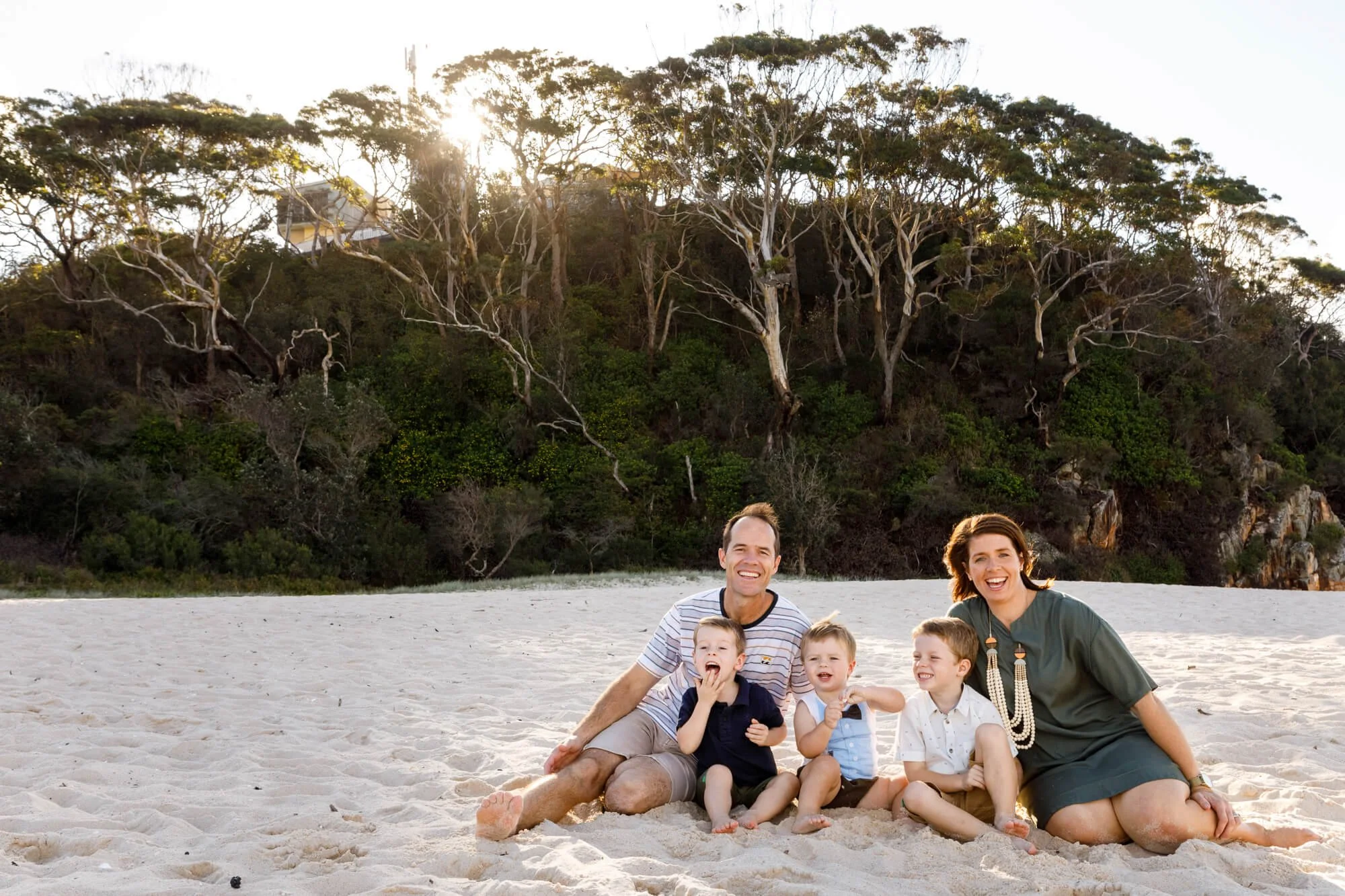 Parents and young children sitting together on the white sand at Shoal Bay with sunlit bushland behind them, Shoal Bay Beach, Nelson Bay, Port Stephens.