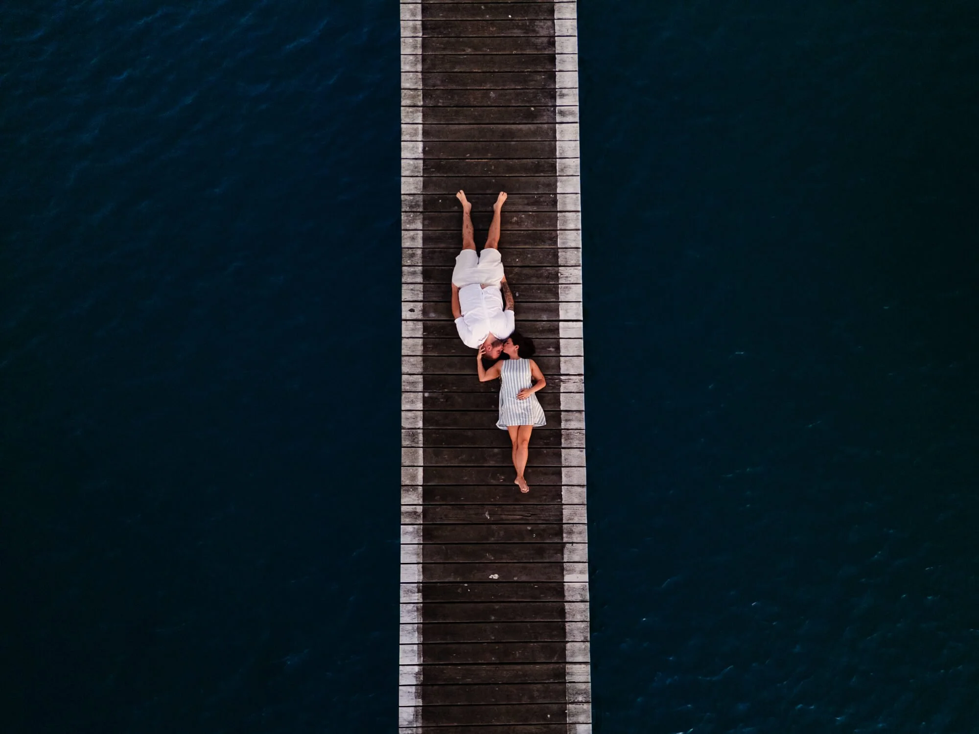 Aerial drone view of a couple lying together at the end of a long timber jetty over dark blue water, Soldiers Point.