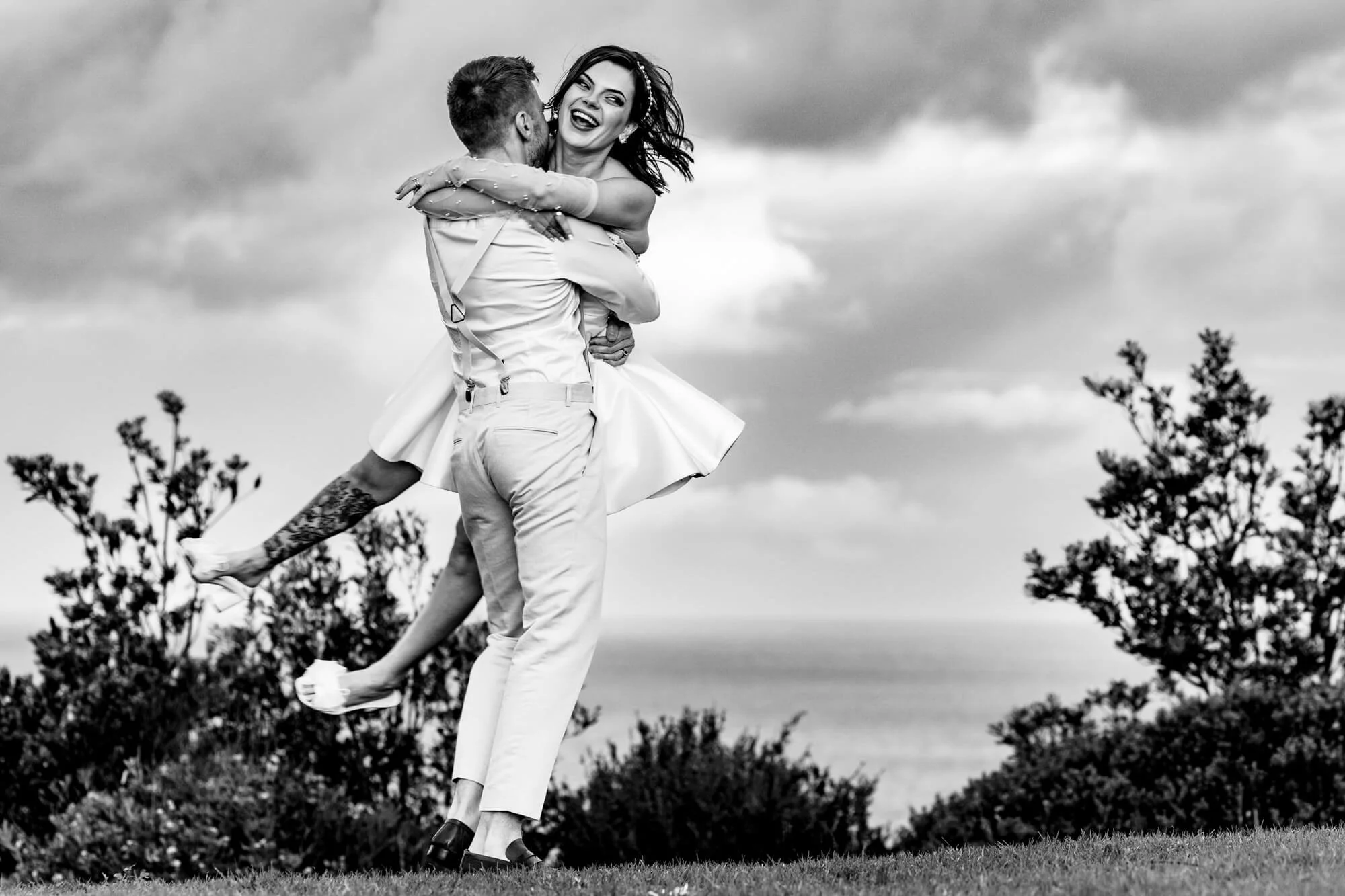 Black and white shot of the groom joyfully lifting his laughing bride off the ground, her shoe kicked loose mid-air, coastal clifftop scrub and ocean behind them.