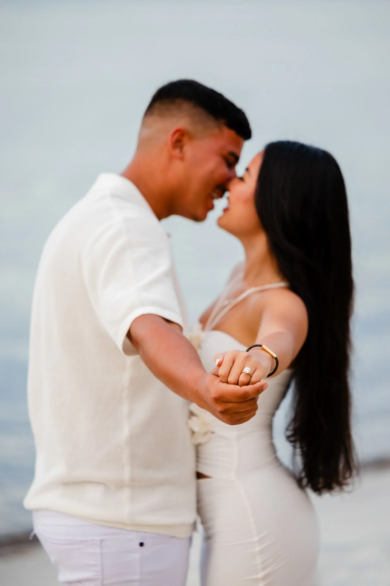 Newly engaged couple almost touching noses on the beach as she shows off her ring, soft blurred background, Dutchmans Beach, Port Stephens.