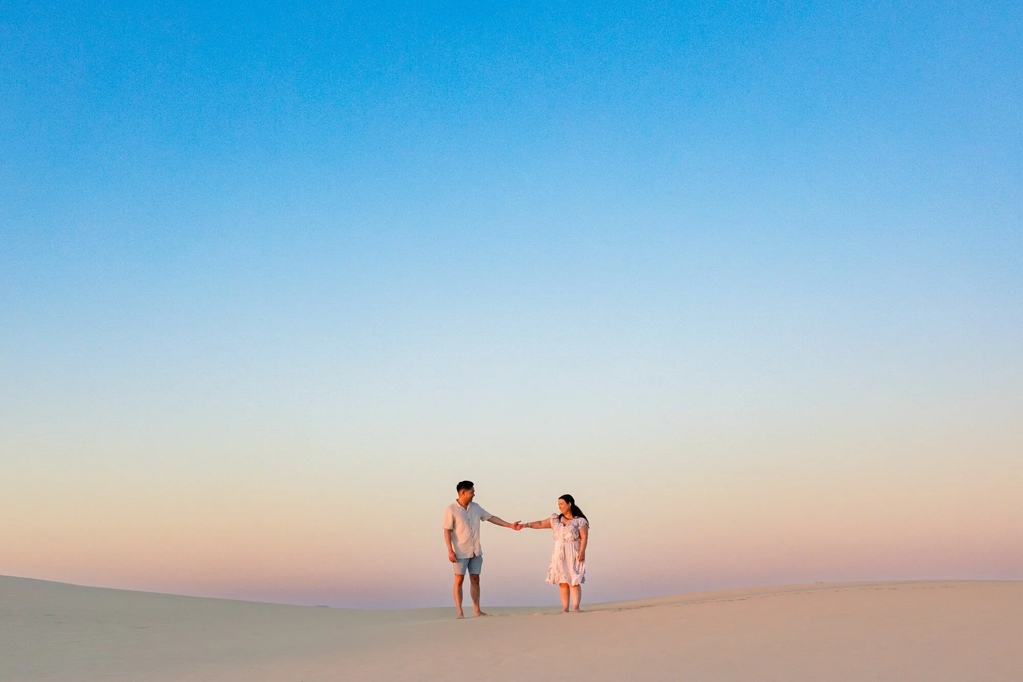 Couple holding hands on a sand dune beneath a soft blue and peach sunset sky, Anna Bay sand dunes, Port Stephens.