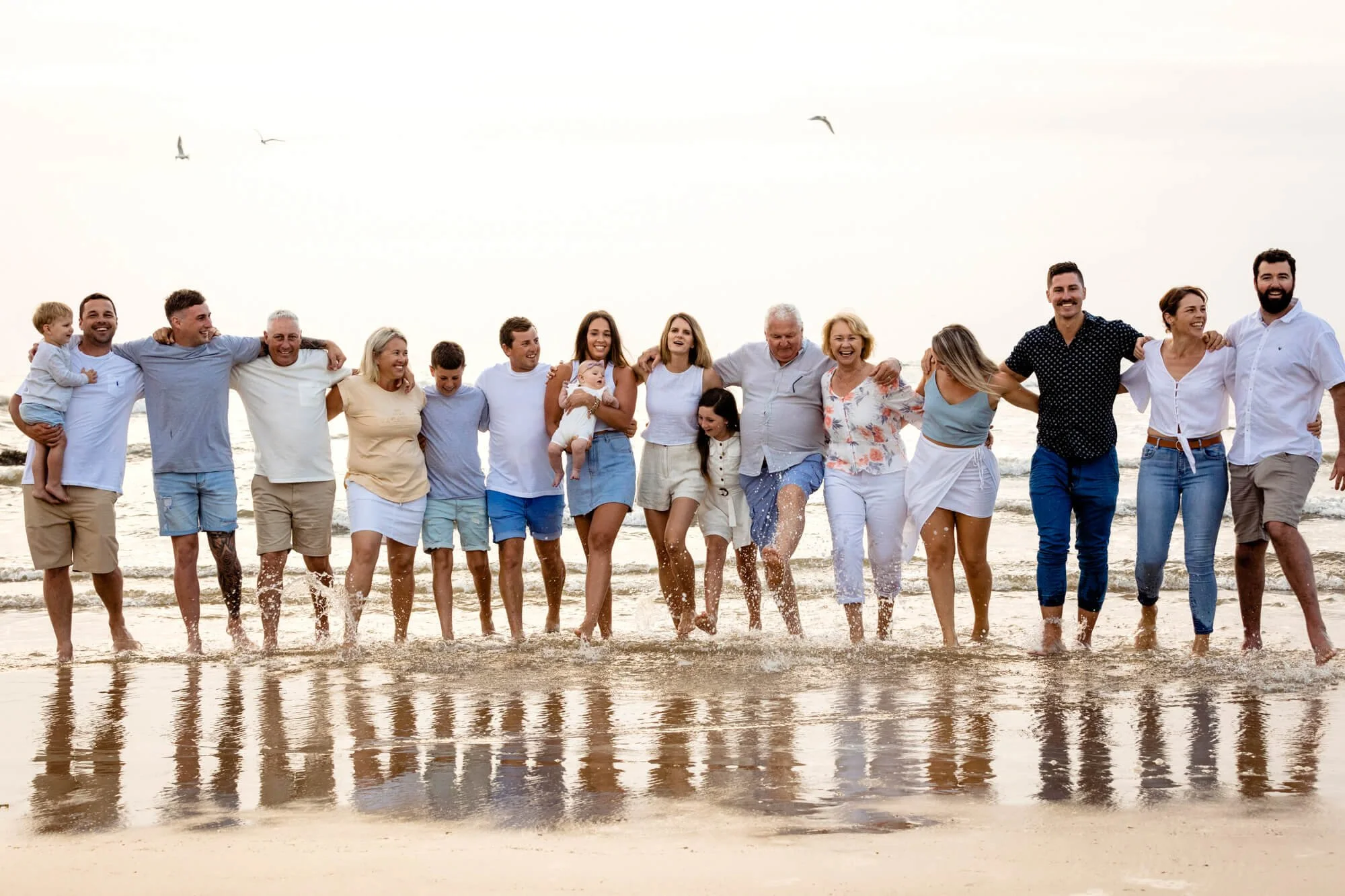 Large extended family standing arm in arm in a long line on a wet beach at sunset, their reflections in the shallow water.