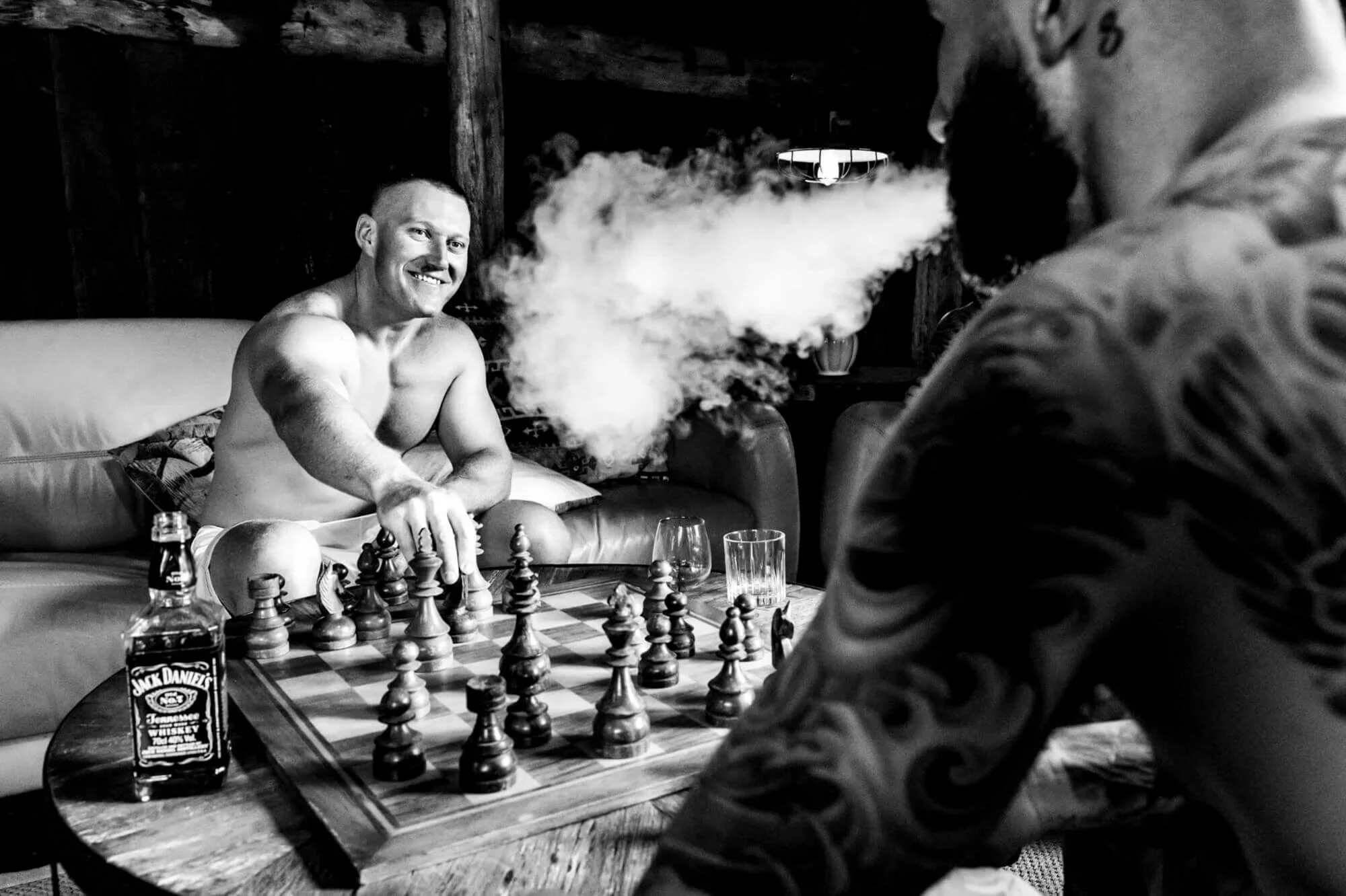 Groom and groomsman playing chess over a bottle of whiskey, cigar smoke curling between them in a relaxed moment before the ceremony.