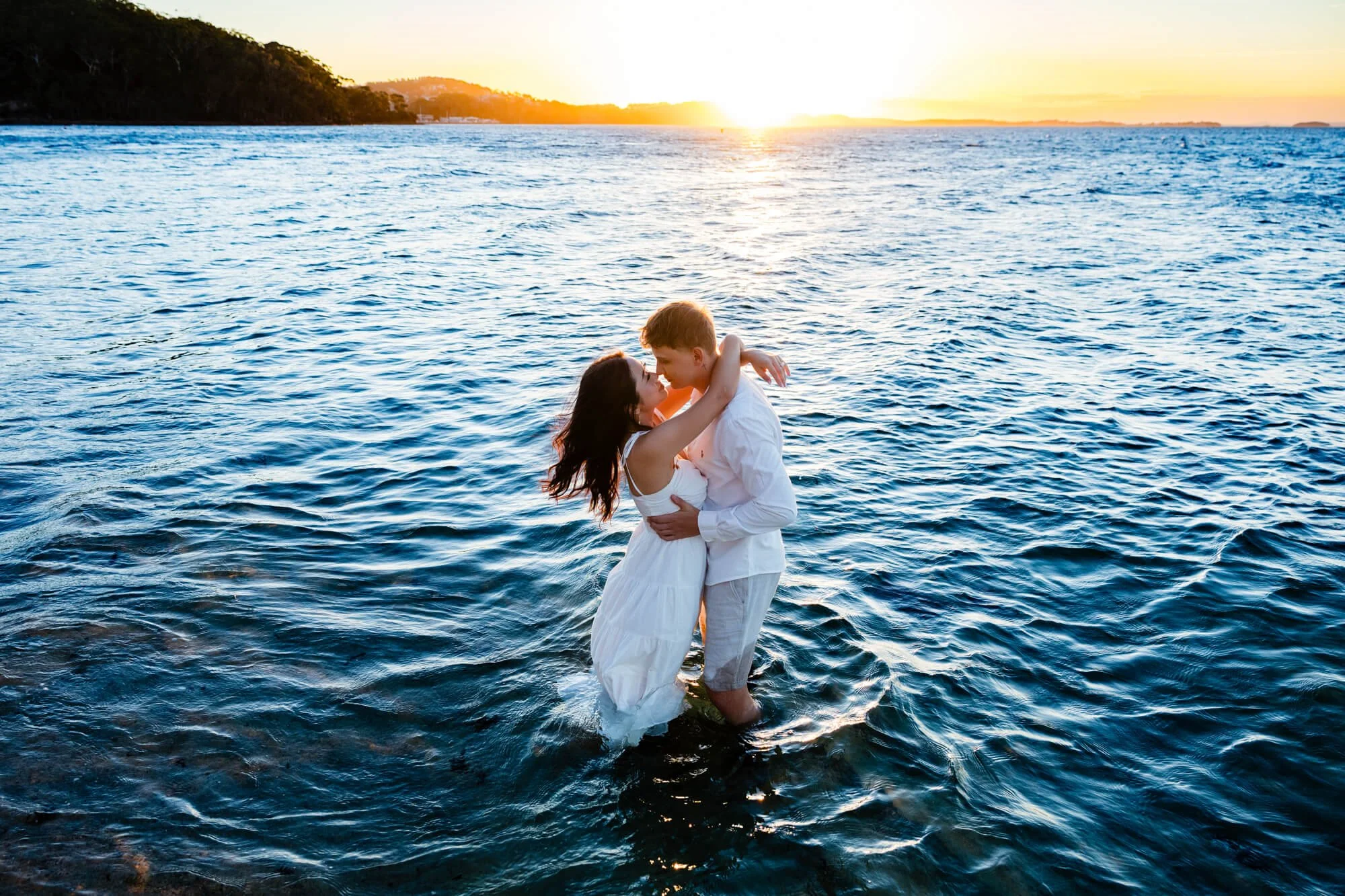 Couple embracing and kissing knee-deep in the water at sunset, the bride's white dress trailing in the ripples, Nelson Bay, Port Stephens.