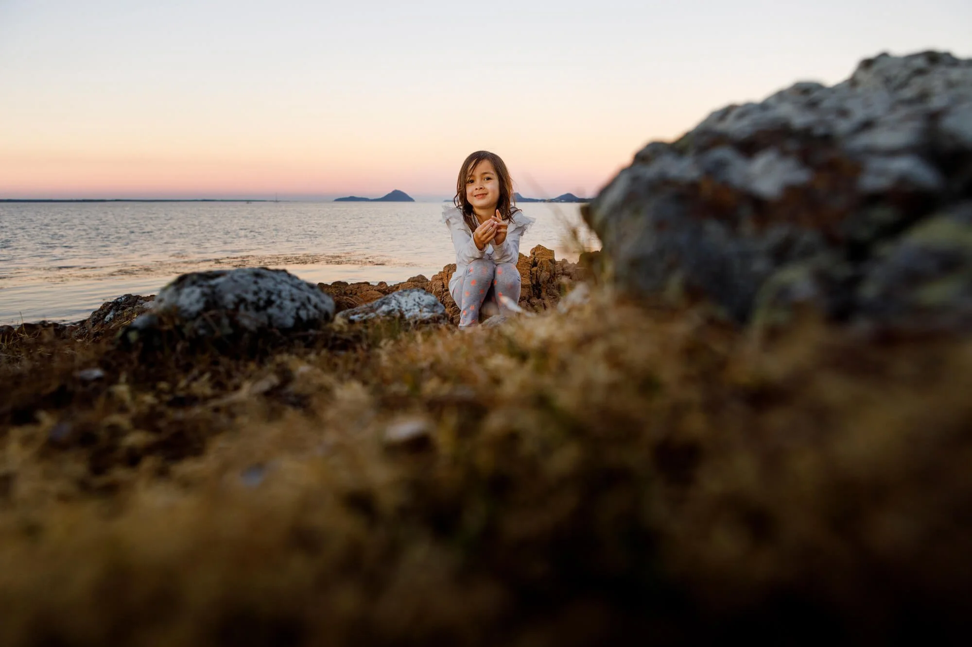 Young child sitting on a rocky shoreline at sunset, foreground grass softly blurred, Soldiers Point, Port Stephens.