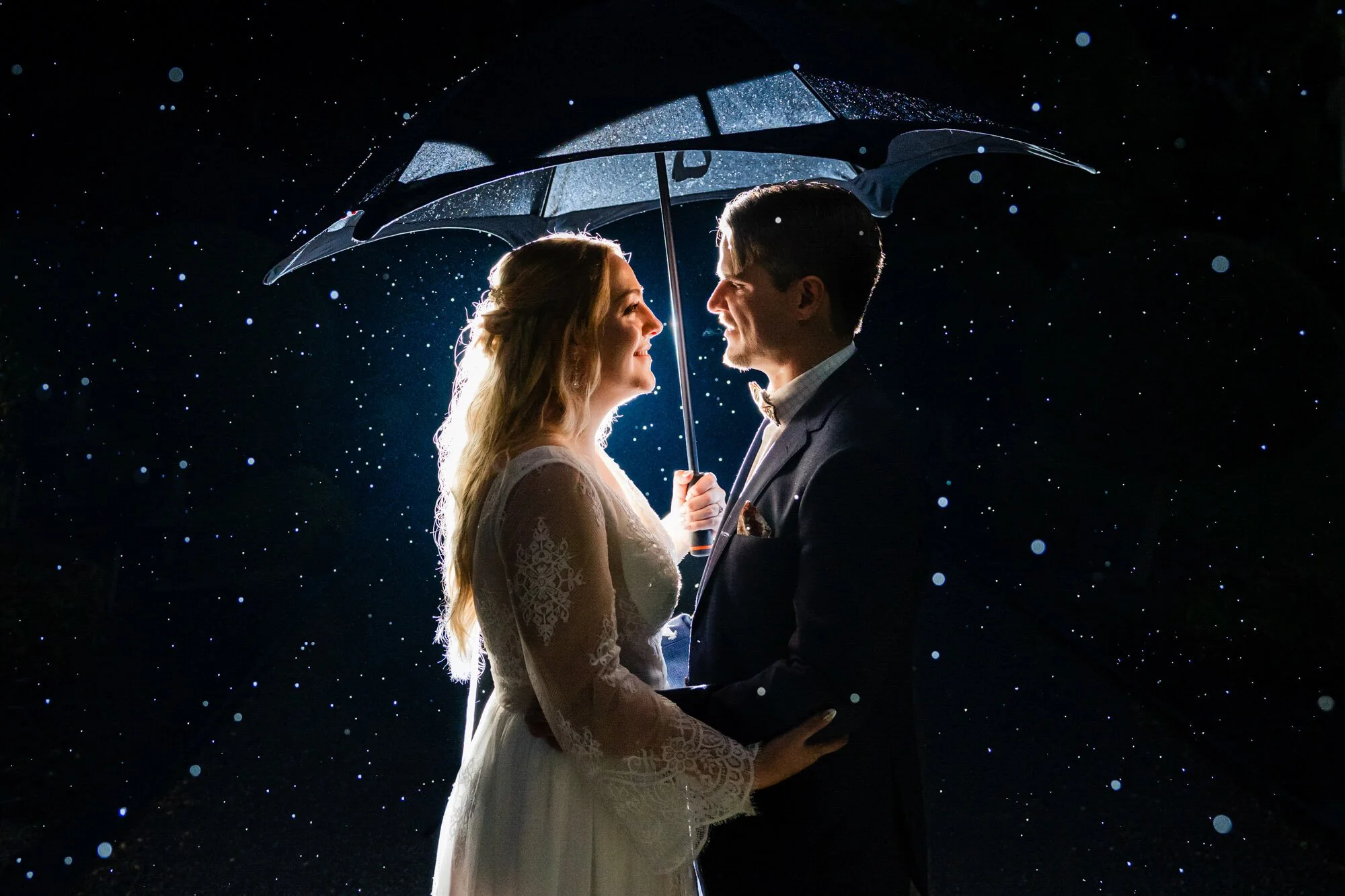 Bride and groom sheltering under an umbrella at night in the rain, droplets sparkling in the flash light.