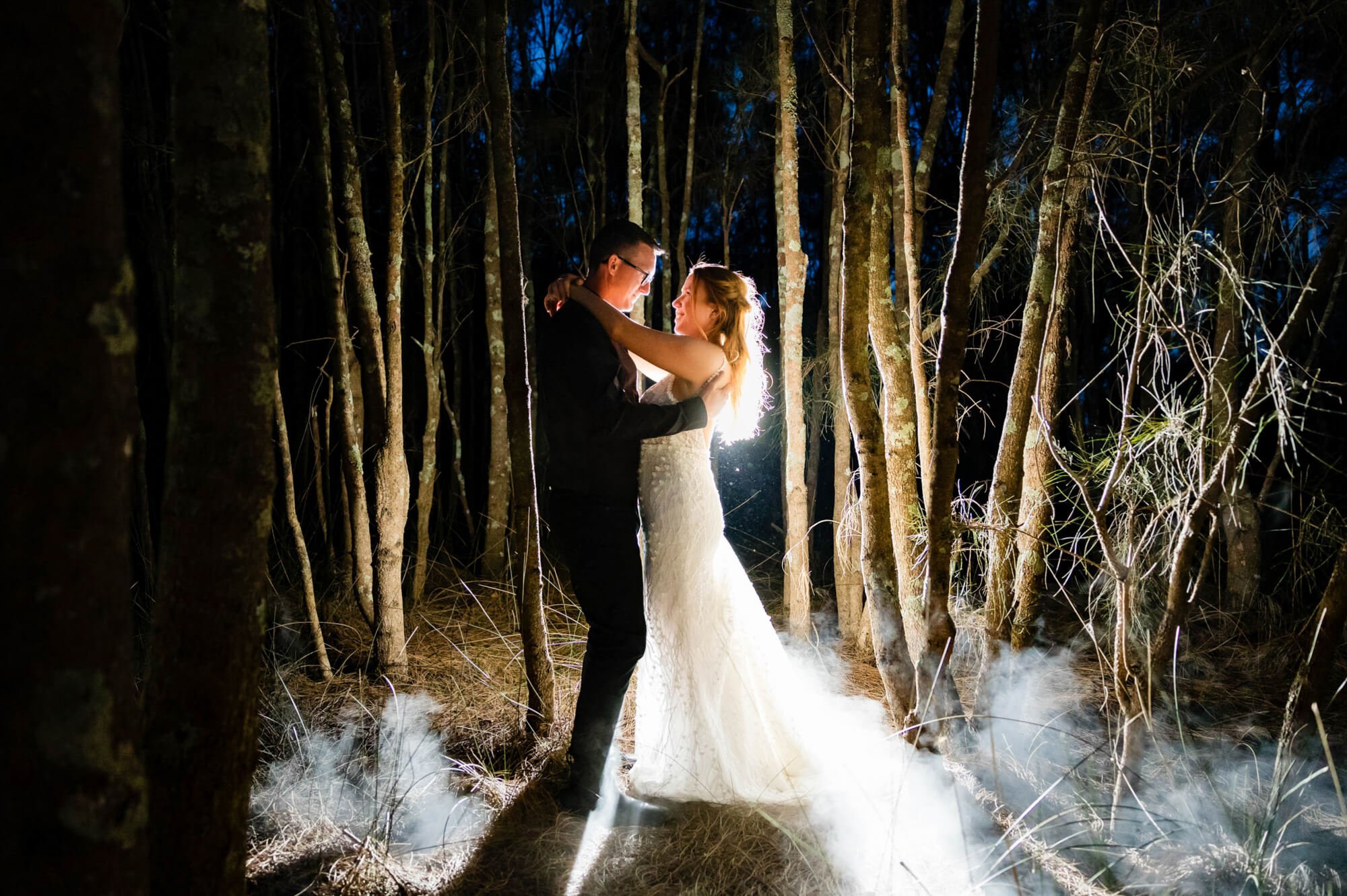Bride and groom sharing an intimate slow dance in a forest clearing at night, warm light glowing through drifting ground smoke among the trees.