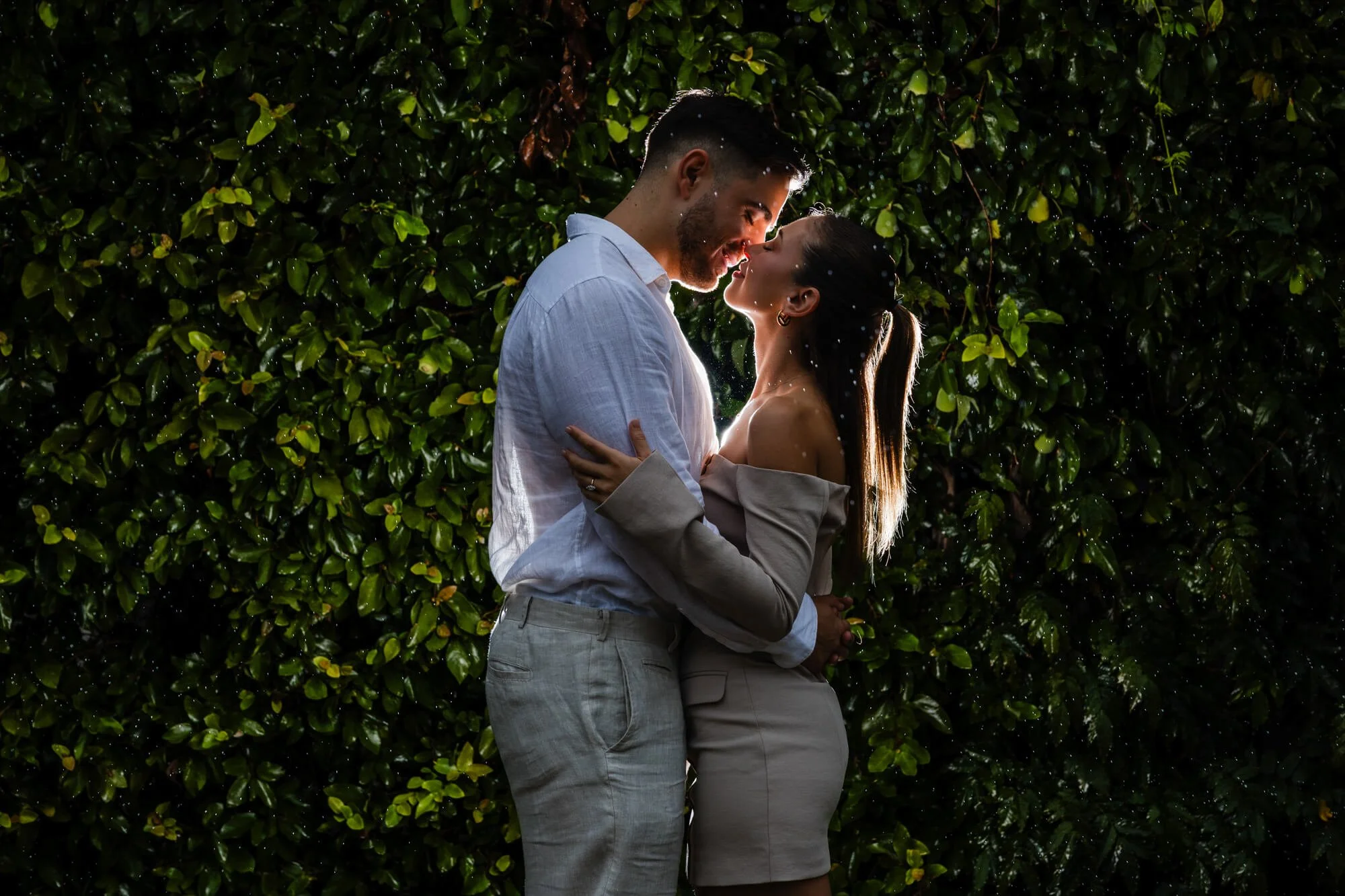 Couple kissing in a warm embrace against a wall of green ivy, soft light catching her hair, just after the proposal, Bob's Farm..