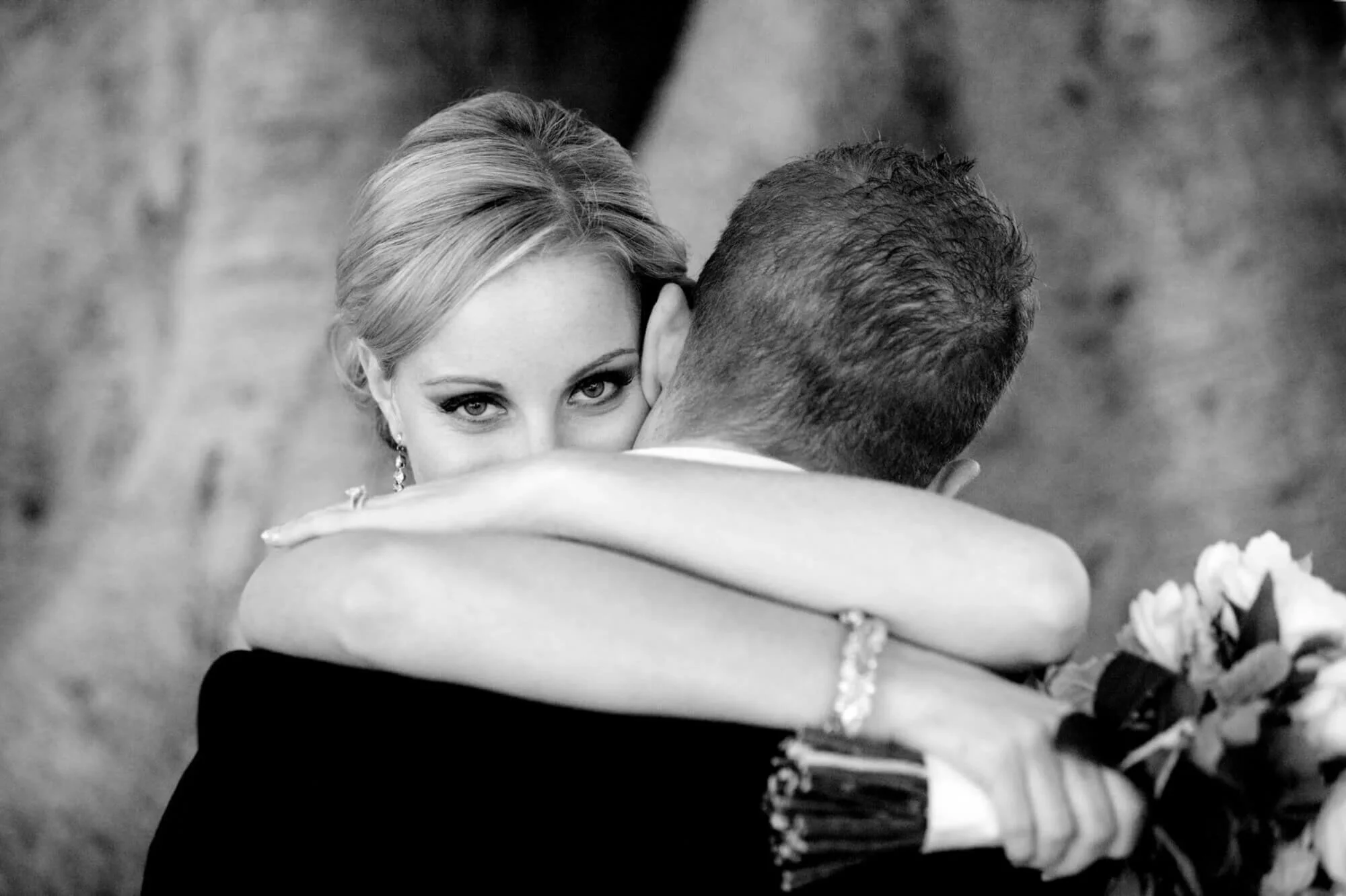 Black and white close-up of the bride resting her chin on her groom's shoulder during a slow dance, her eyes looking straight at the camera, bouquet still in hand.
