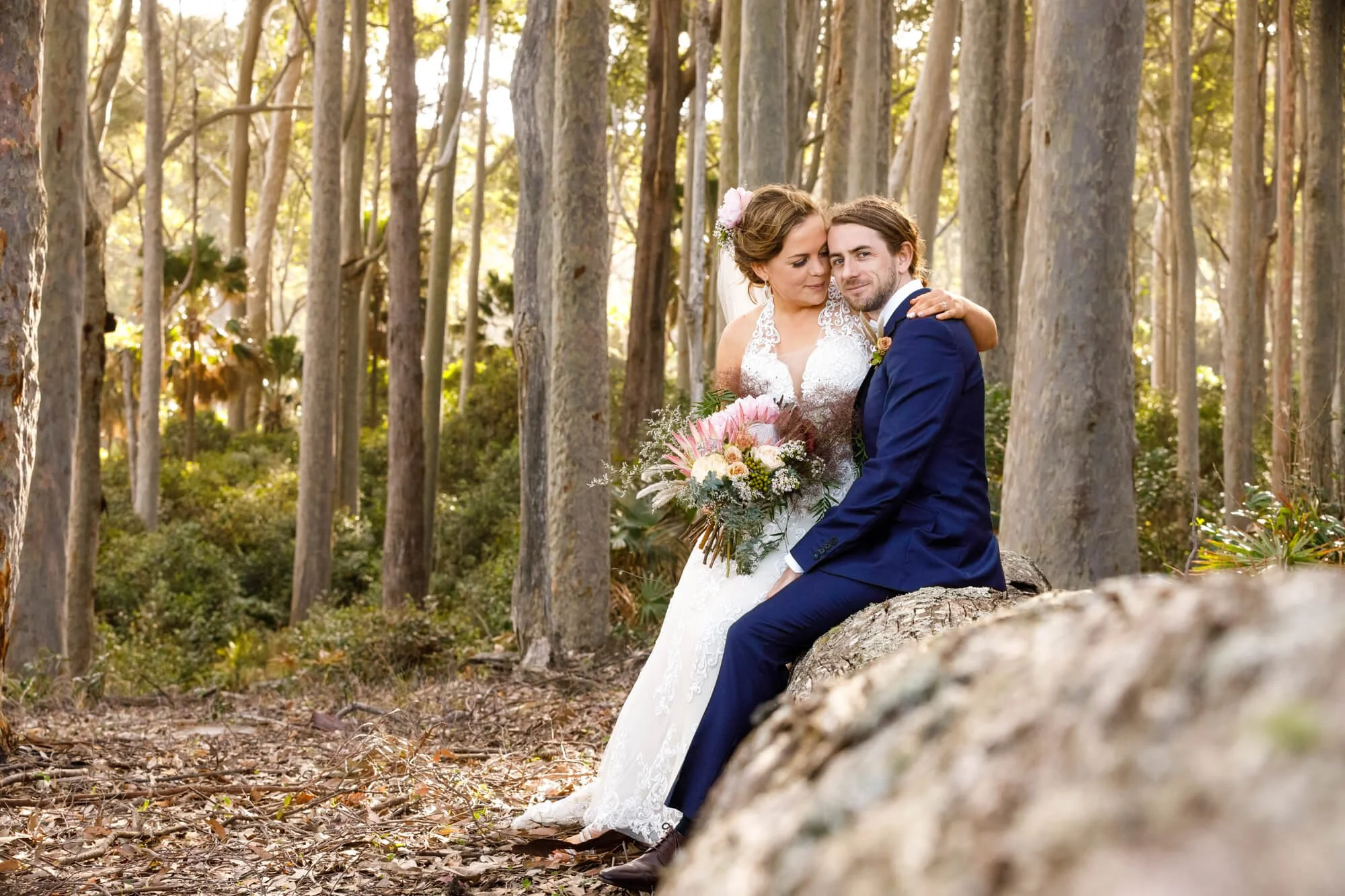 Bride and groom sitting together on a rock in the forest, her bouquet in hand, surrounded by tall trees.