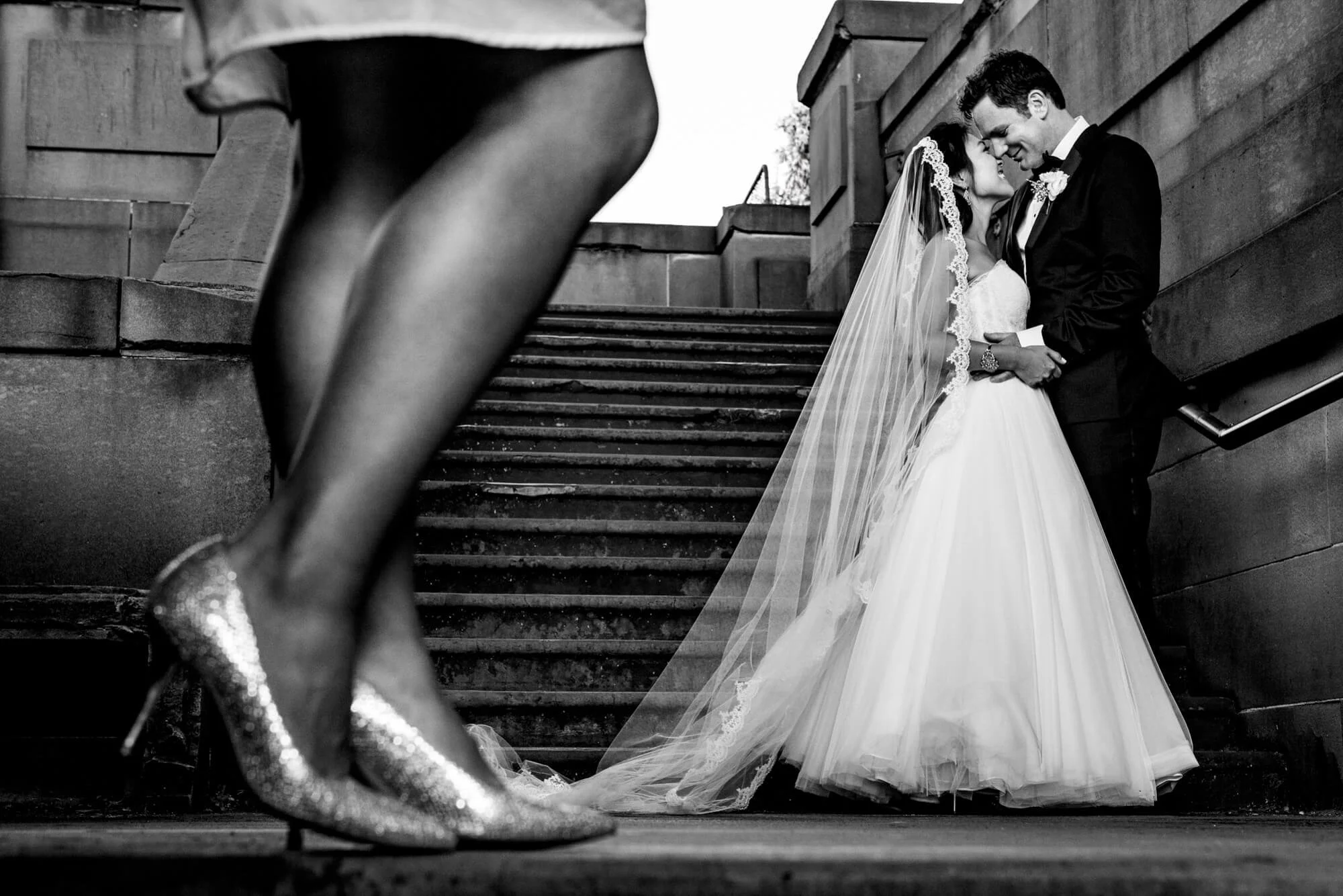 Creative black-and-white composition layering a bridesmaid's sparkly heels in the foreground with the bride and groom embracing on stone stairs behind, The Rocks, Sydney