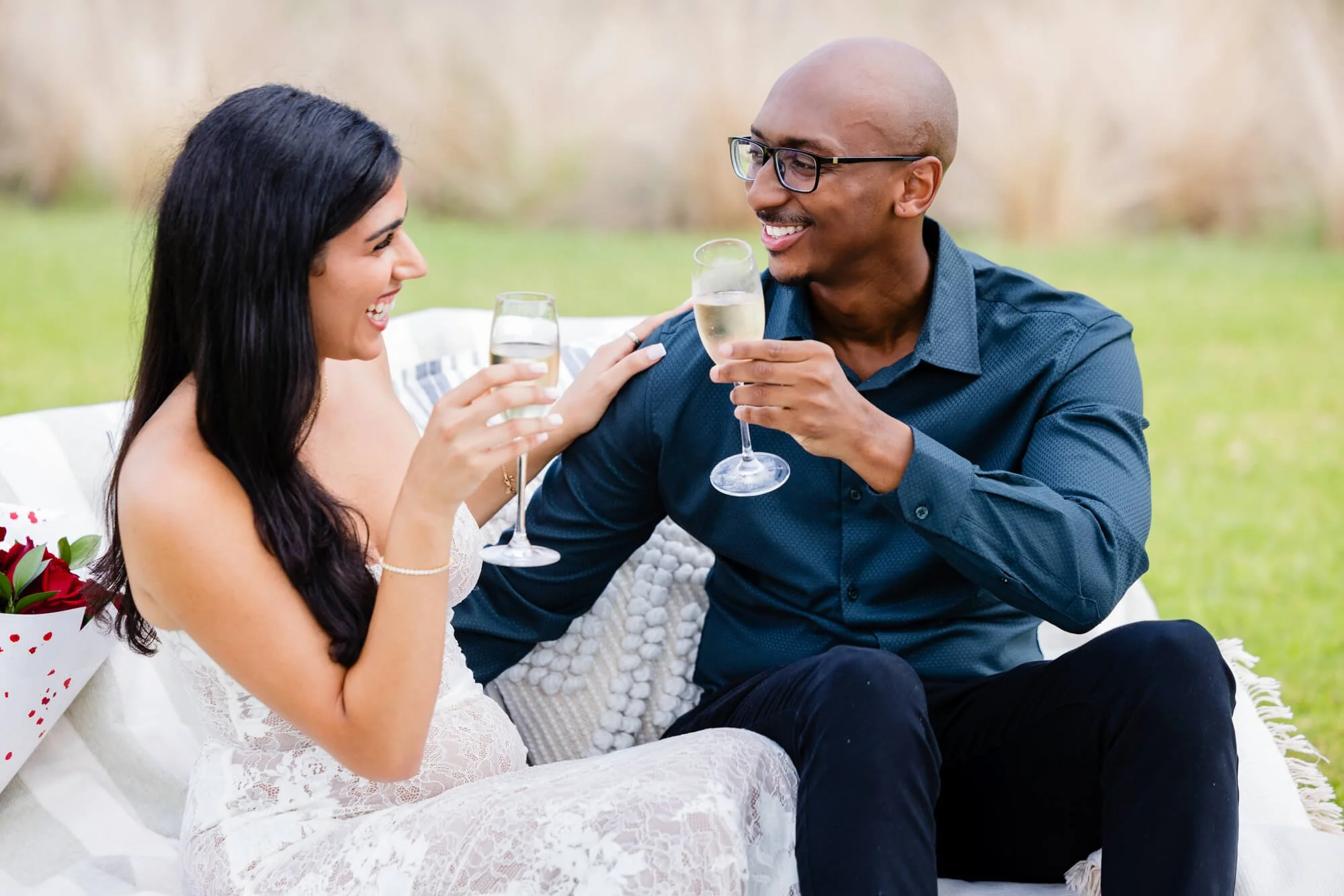 Couple laughing together and raising champagne flutes on a white picnic rug in a grassy field, Hunter Valley.