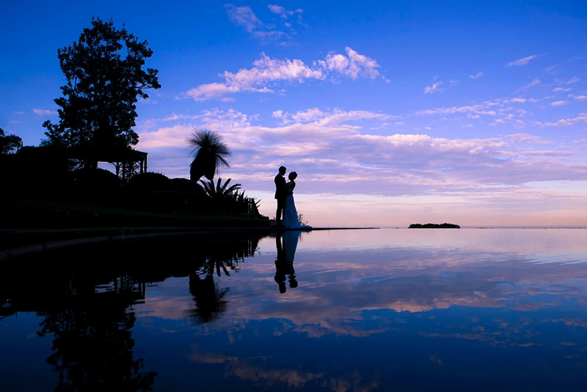 Bride and groom silhouetted against a dusky blue sky, their reflection mirrored perfectly in the still water below.
