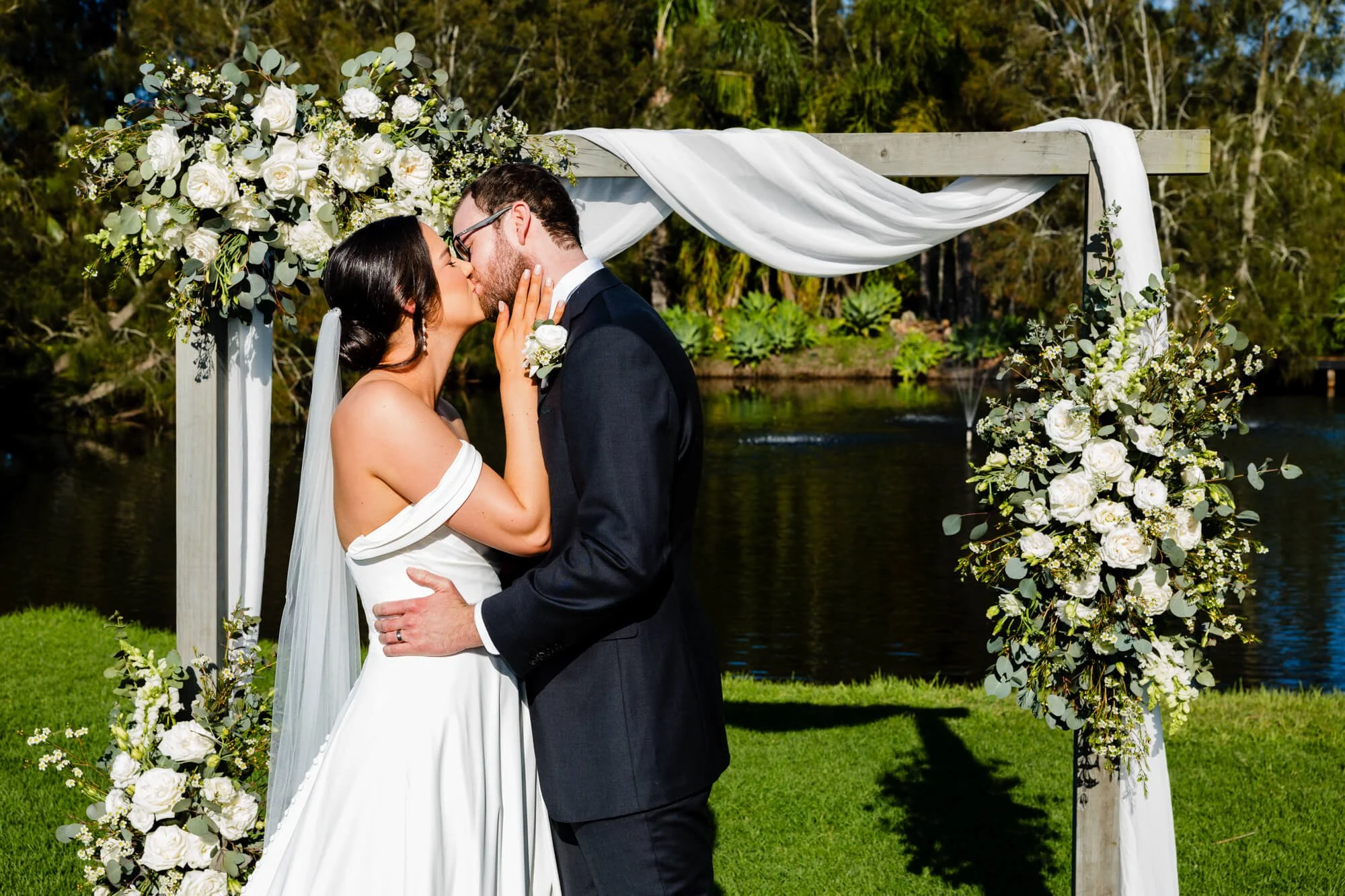 Bride and groom kissing under a white floral arbor beside the water during their elopement ceremony.