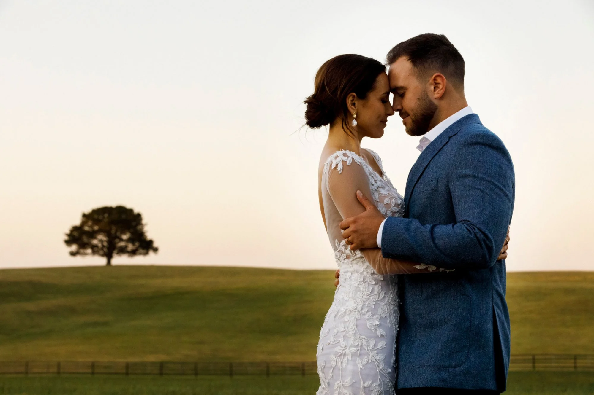 Bride and groom standing forehead to forehead in an open paddock at sunset, a single tree silhouetted in the distance.
