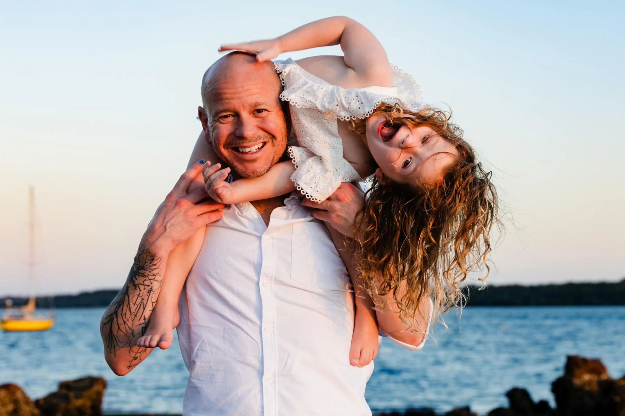 Dad laughing with his daughter on his shoulders by the water at golden hour, boats on the bay behind them, Lemon Tree Passage, Port Stephens.