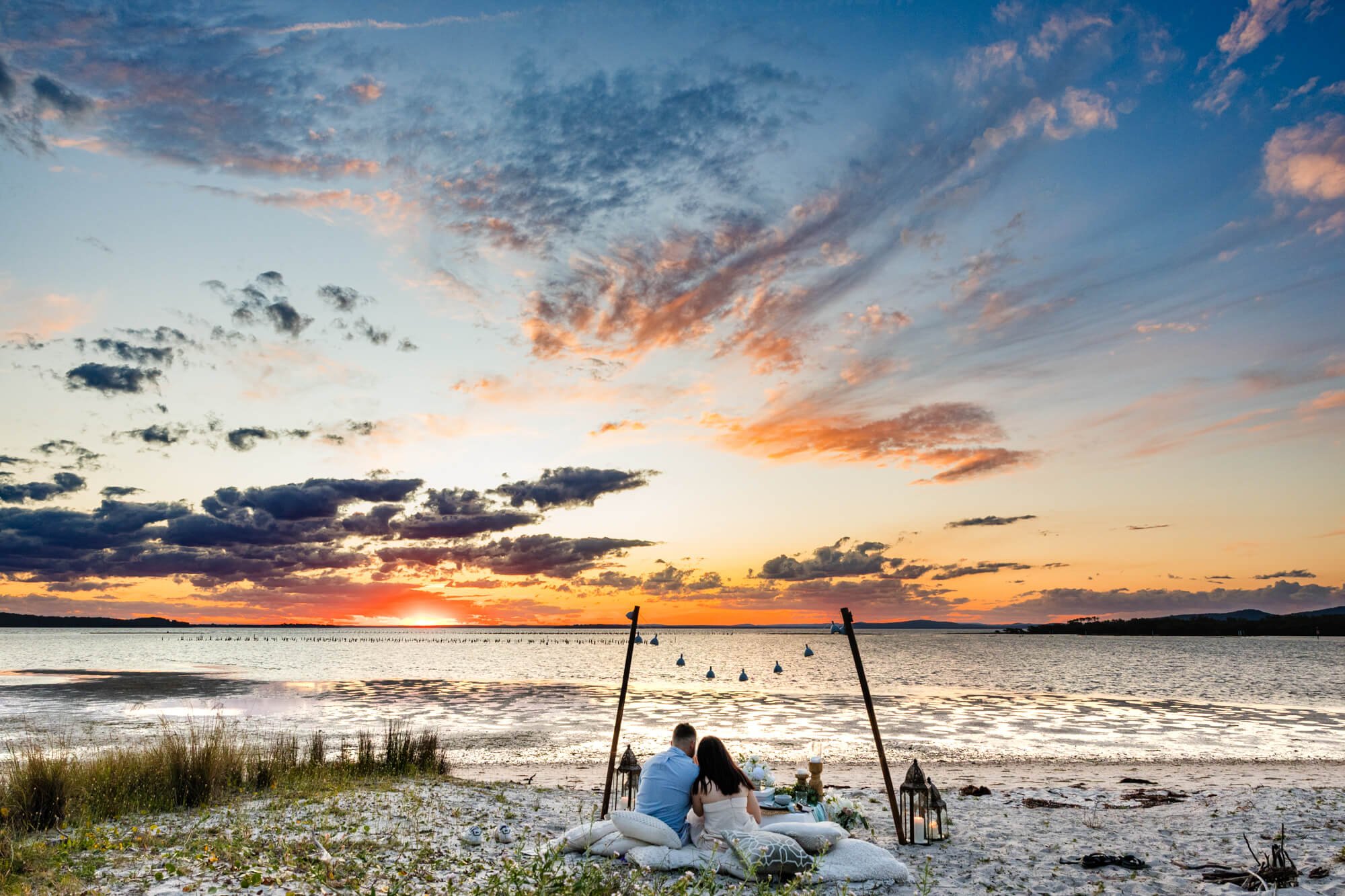 Couple sitting together on cushions at a styled beach picnic under a vivid orange and blue sunset sky, lanterns and poles marking out the setup, Soldiers Point.