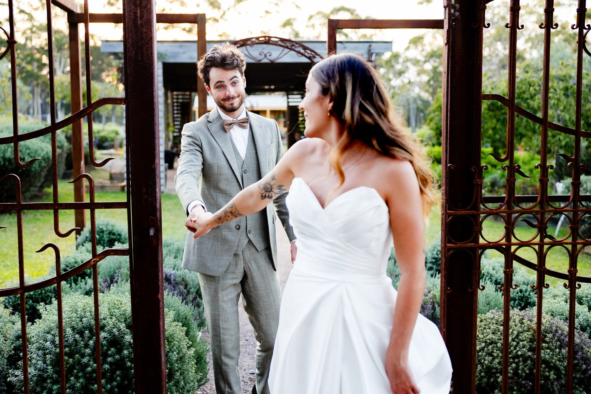 Groom reaching for his bride's hand as she walks toward him through a weathered iron garden gate.