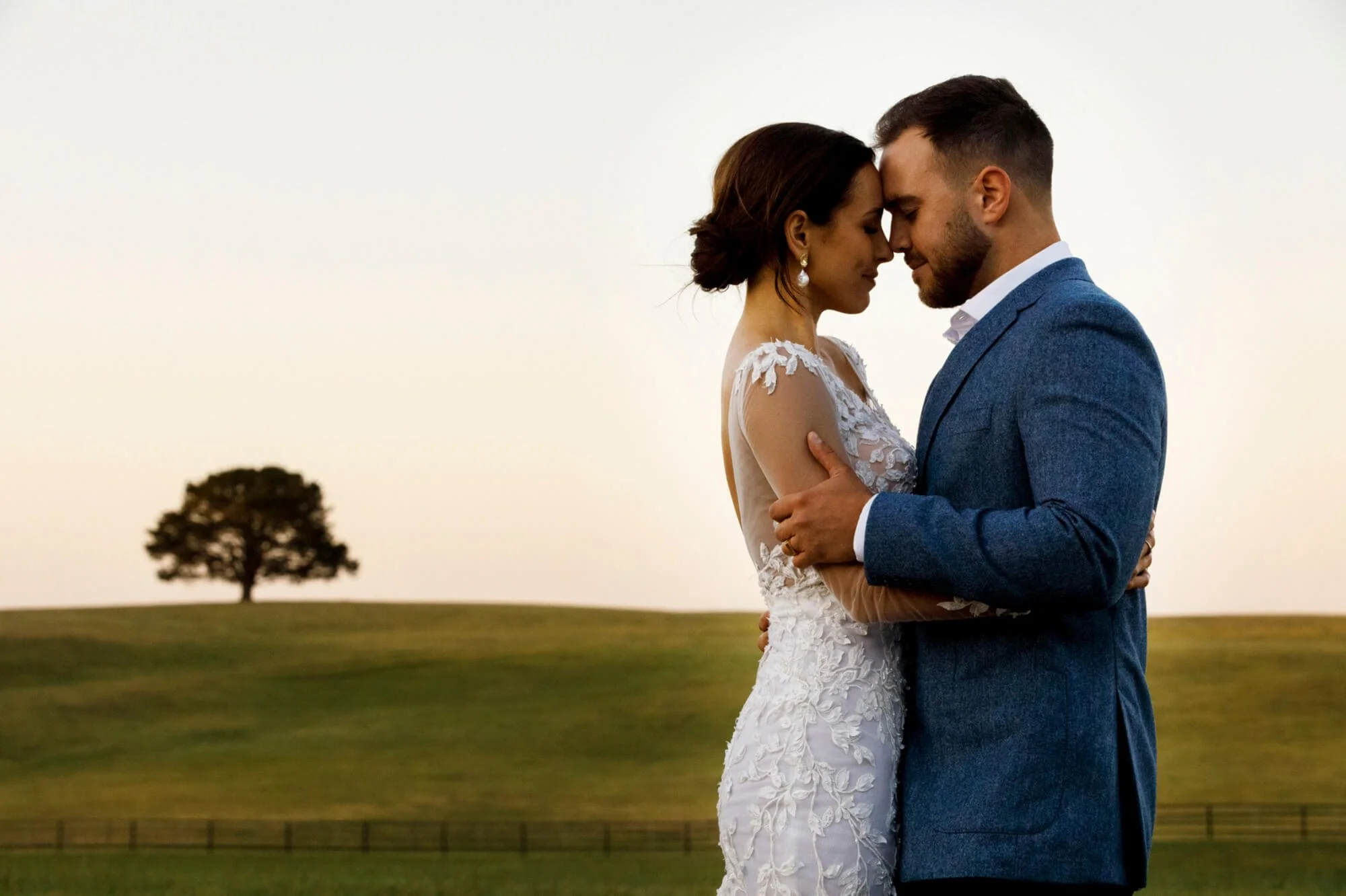 Bride and groom forehead to forehead in an intimate embrace, a lone tree and rolling green paddocks behind them at dusk.