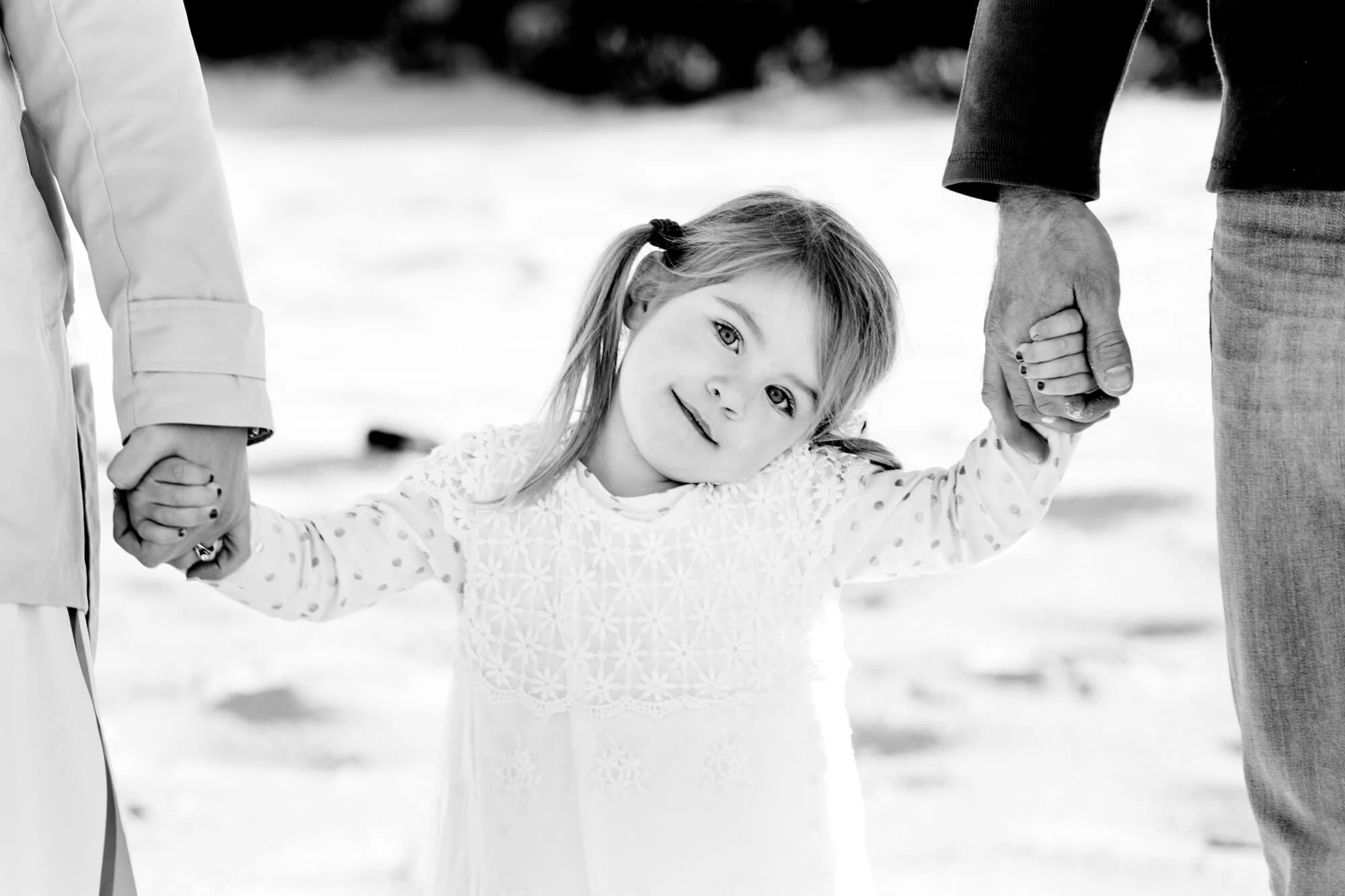 Black and white portrait of a little girl smiling at the camera while holding her parents' hands on the beach, Shoal Bay Beach, Nelson Bay, Port Stephens.