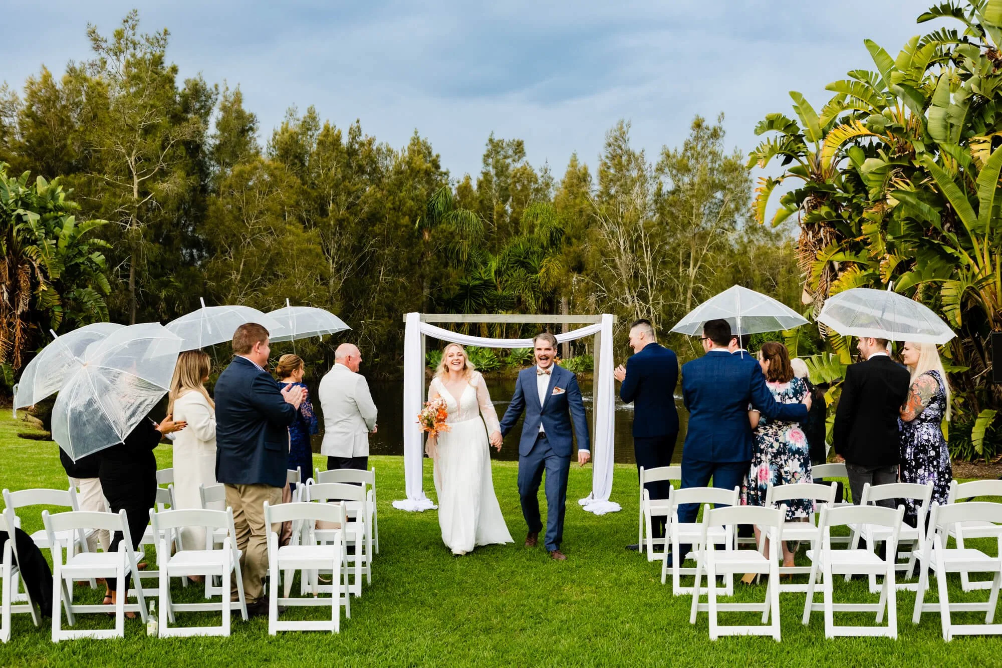 Small elopement ceremony at Heaven Gardens with the couple exchanging vows under clear umbrellas as guests look on from white chairs under an overcast sky.
