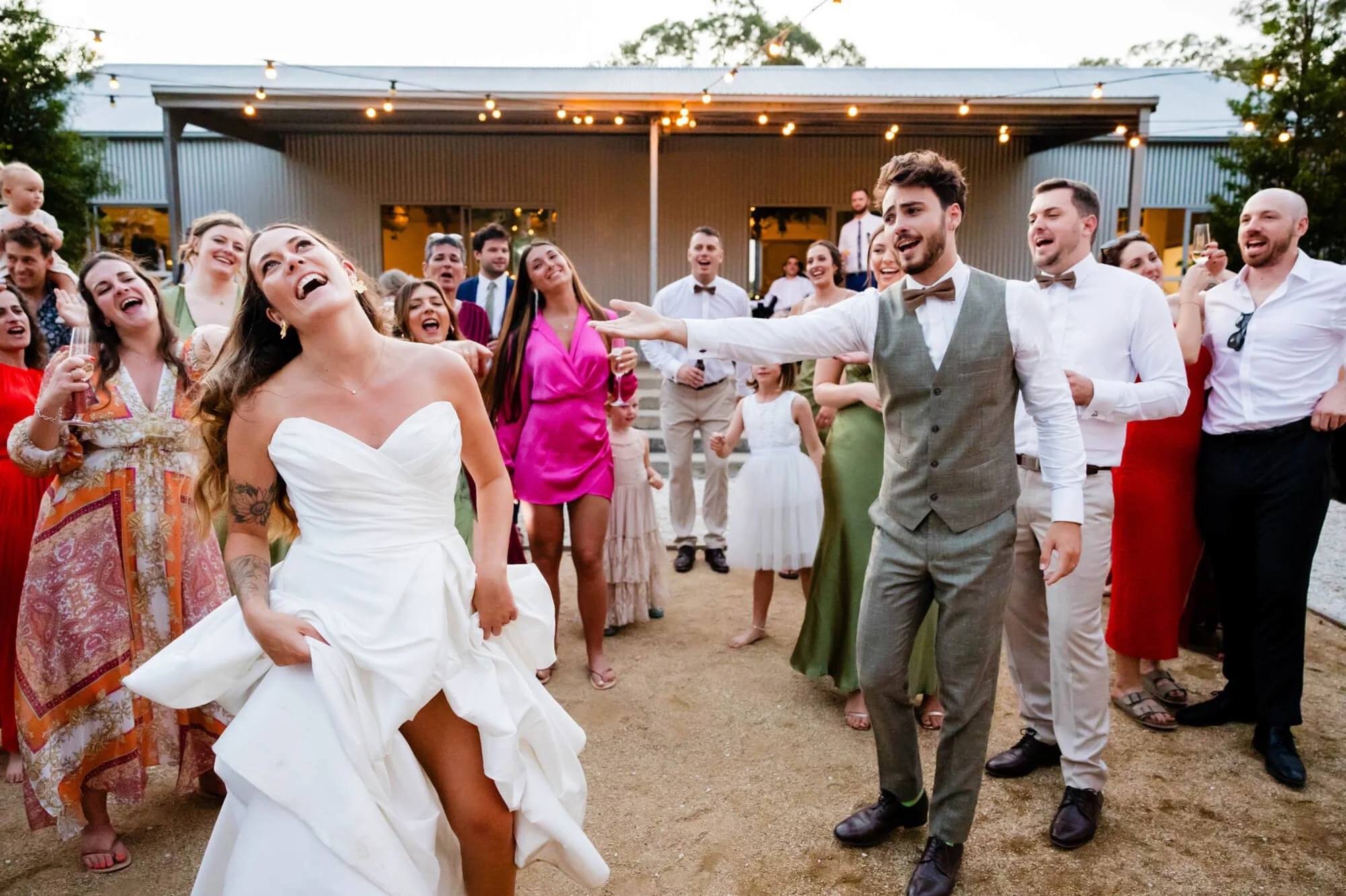 Bride and groom laughing and dancing together as guests cheer them on under festoon lights at the reception venue.