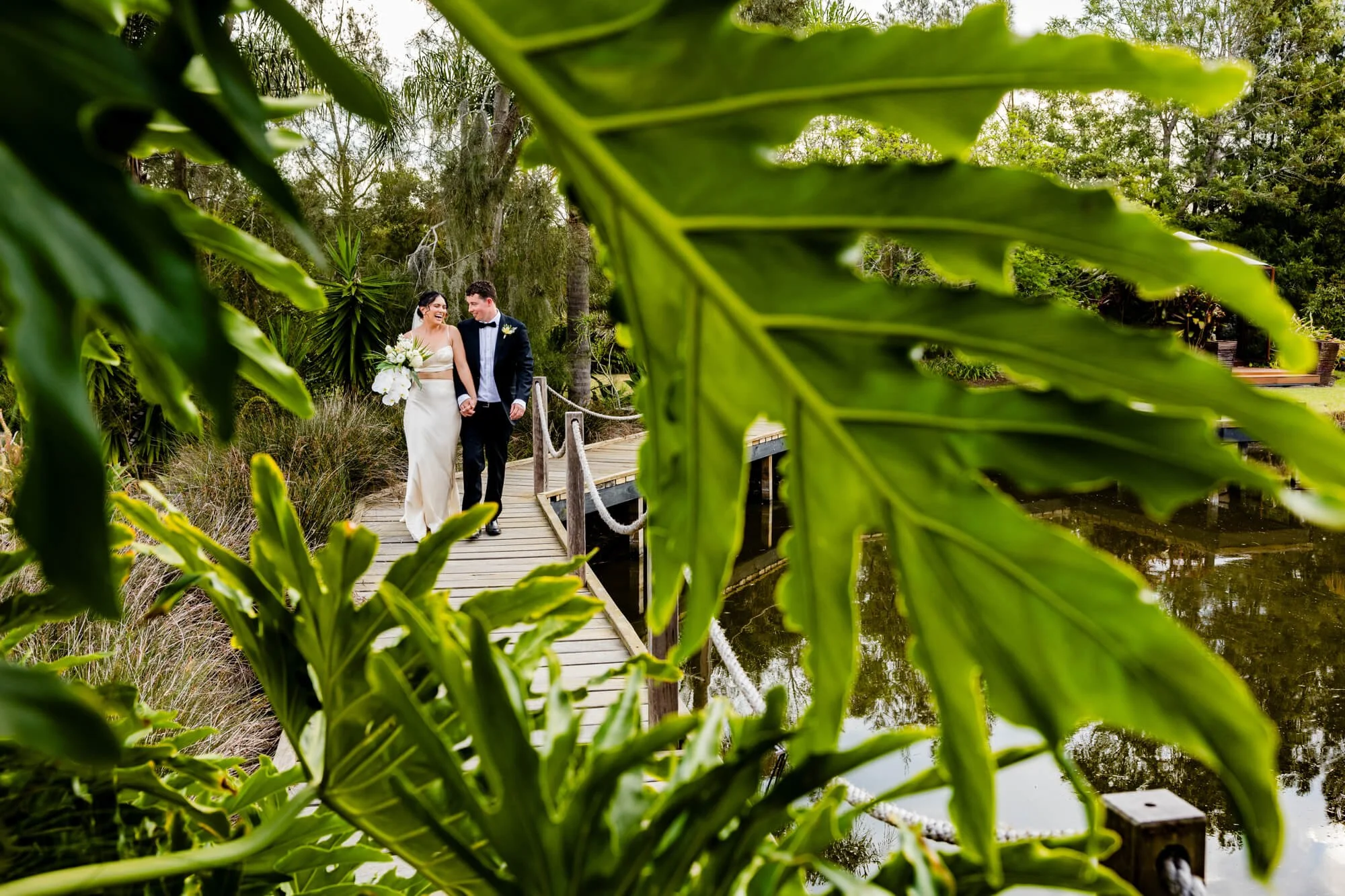 Bride and groom walk hand in hand along a fern-lined path through the lush greenery of Heaven Gardens, Salt Ash, Port Stephens.