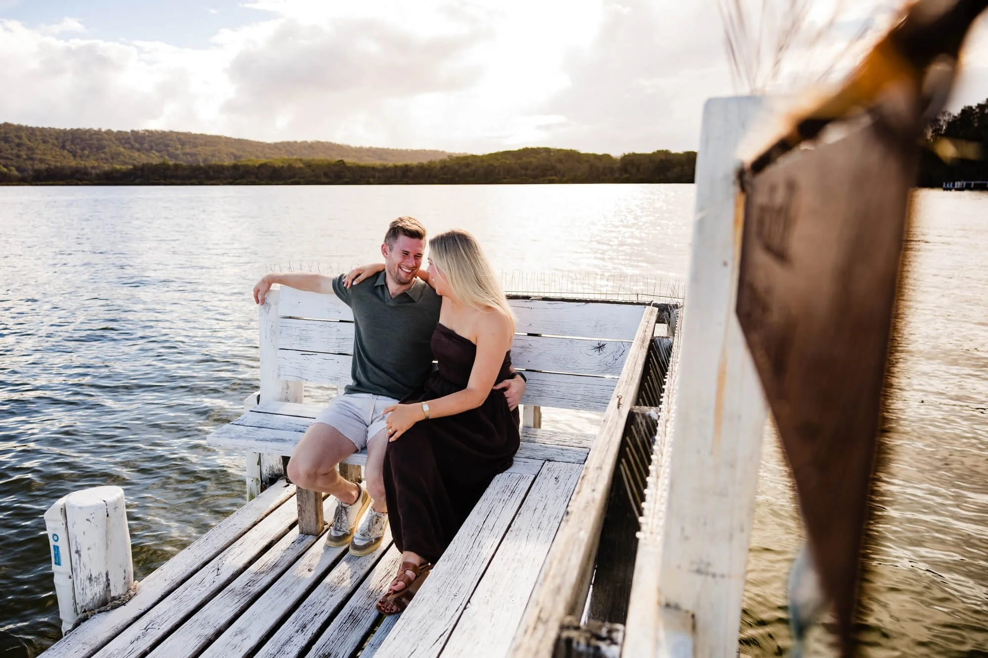 Couple sitting close together on a weathered white timber jetty seat over calm water at dusk, a wooded shoreline behind them, Mid North Coast.