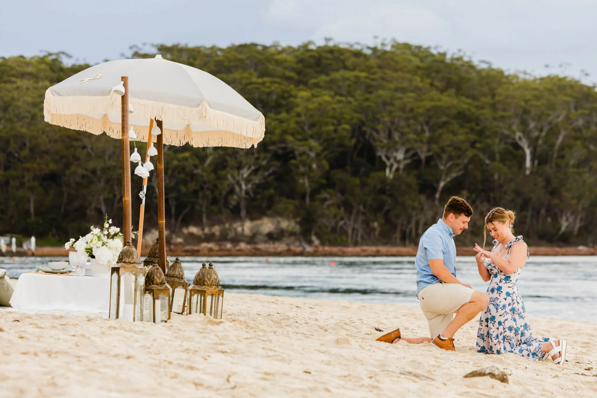 Man down on one knee proposing to his partner on a white-sand beach beside a fringed cream umbrella and lanterns, bushland and calm water behind them, Little Beach.