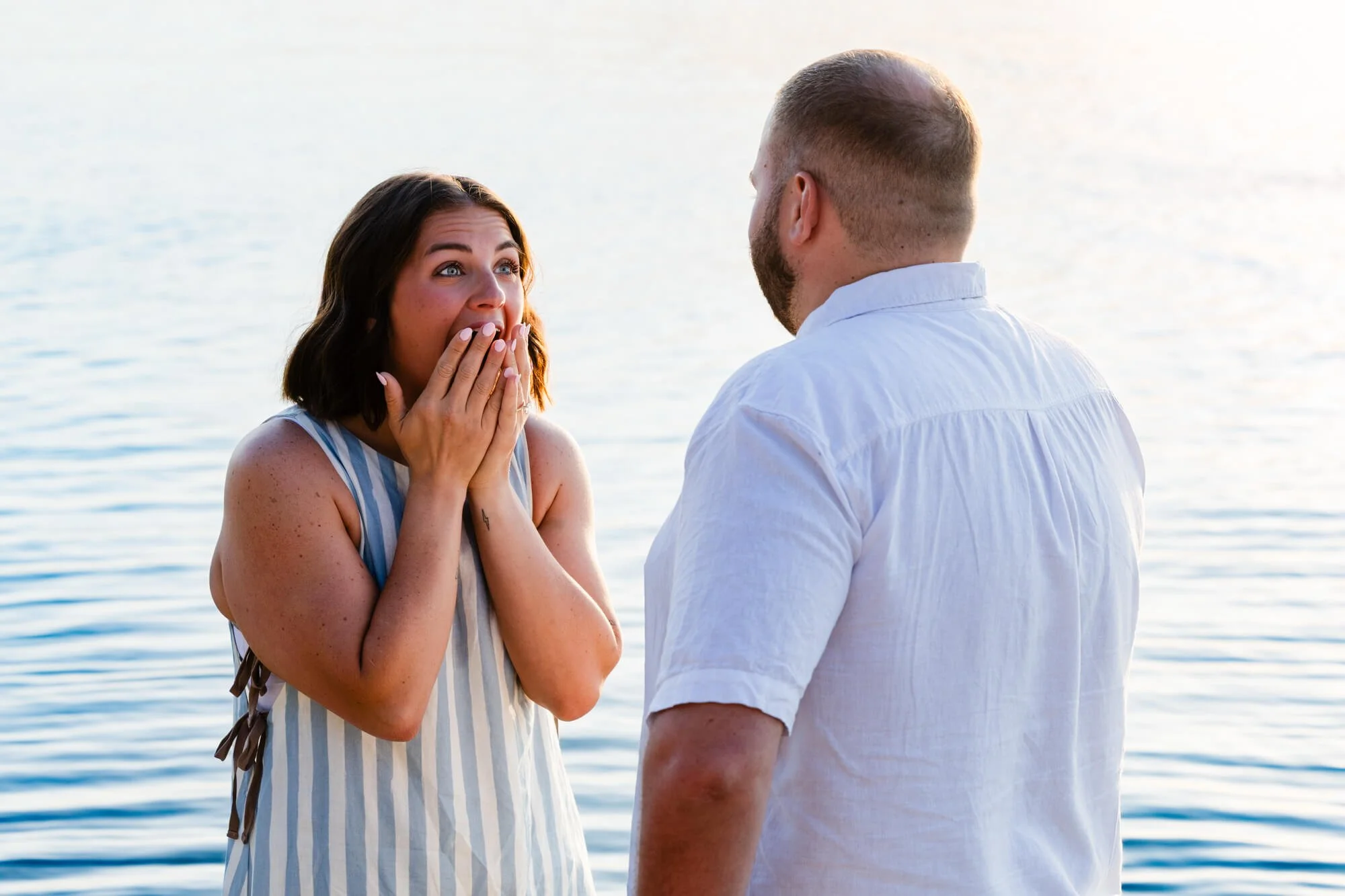 Woman with her hands over her mouth in happy shock as her partner faces her at the water's edge, a candid reaction moments after the proposal, Soldiers Point.