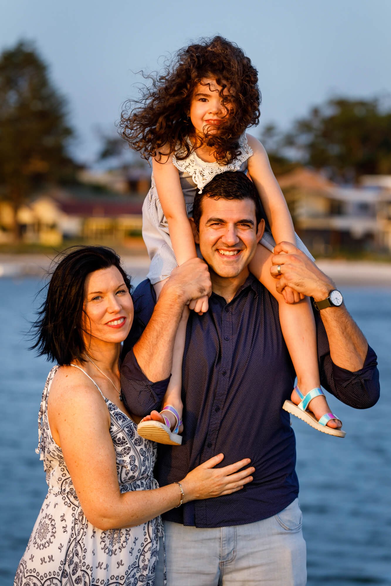 Mum and dad standing in the shallows at sunset with their daughter on dad's shoulders, Soldiers Point, Port Stephens.