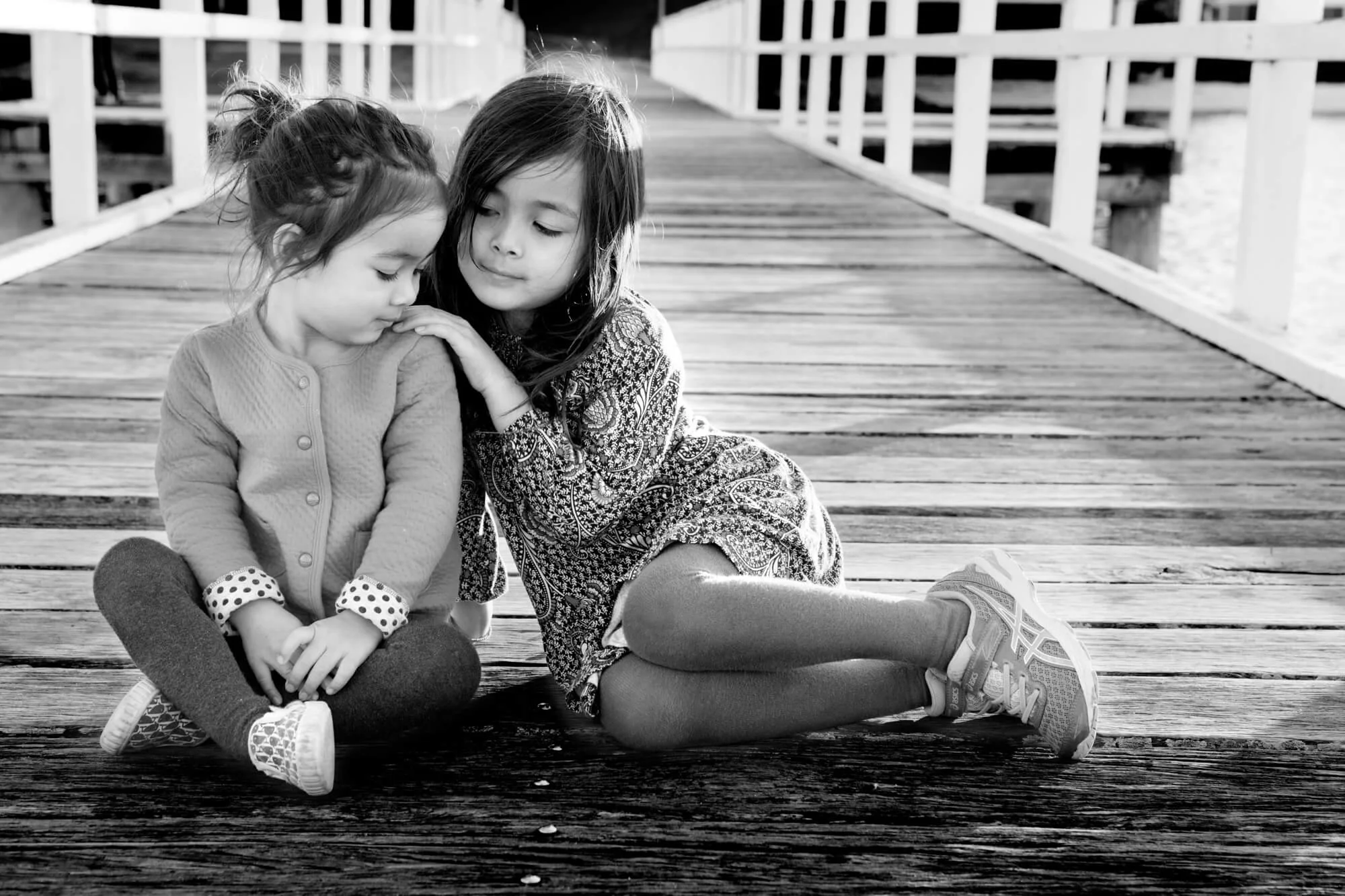 Black and white photo of two sisters sitting close together on an old timber jetty, one resting her head on the other's shoulder, Salamander Bay, Port Stephens.