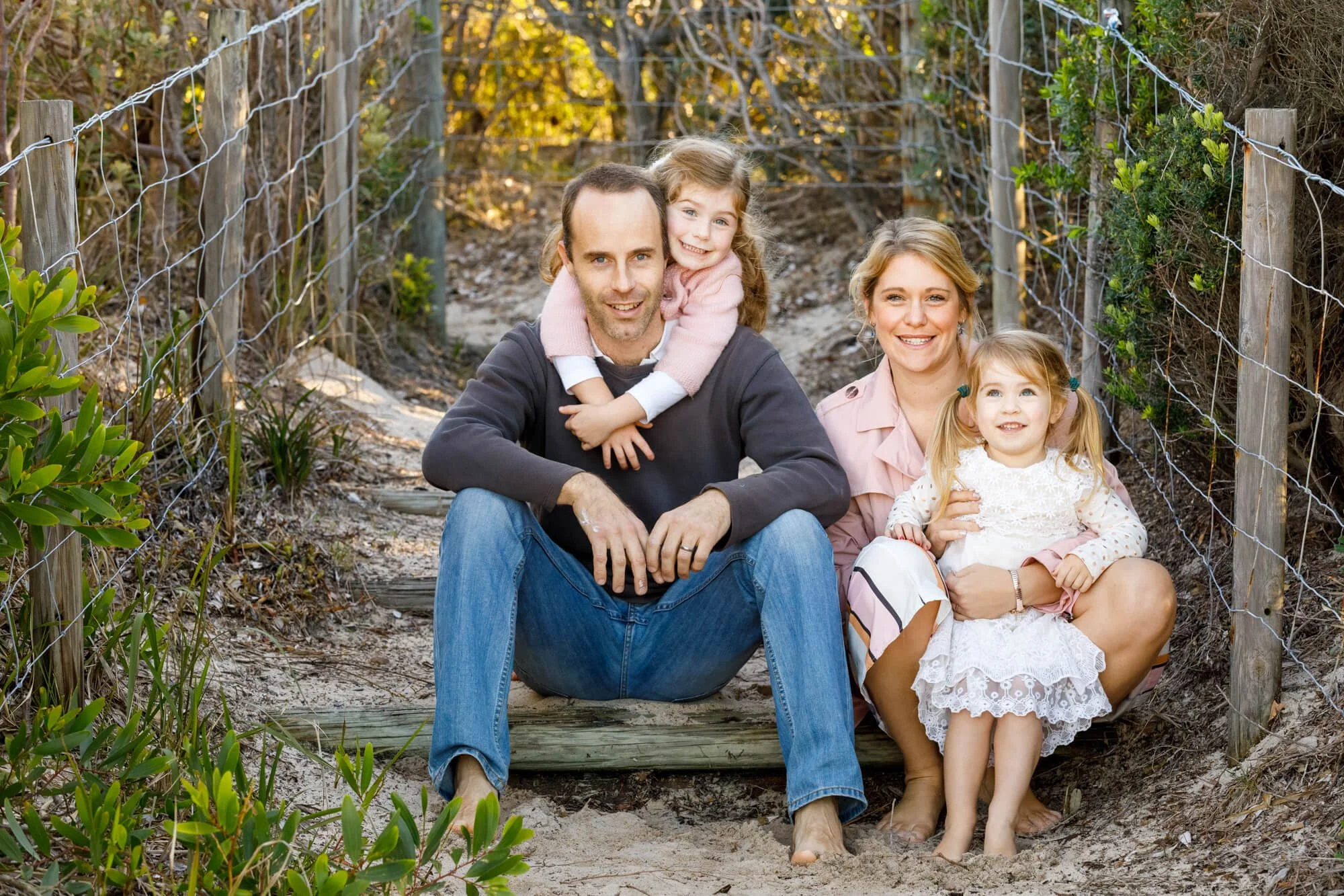 Mum and dad sitting on a timber step beside a fence on a bush track with their two young daughters, Bagnalls Beach, Corlette, Port Stephens.