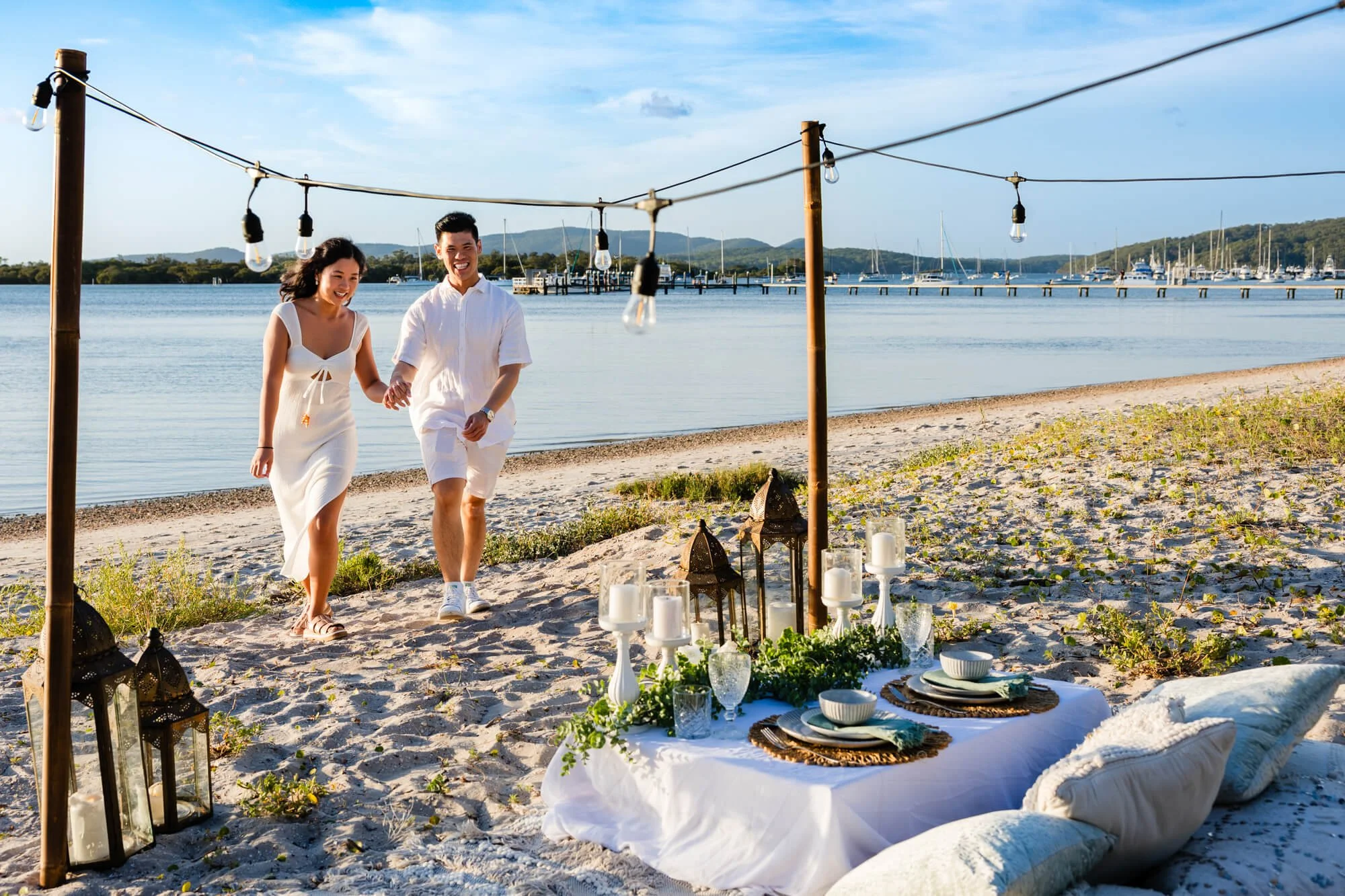 Couple in white walking hand in hand along the beach towards a styled picnic table with lanterns, string lights and cushions, Soldiers Point, Port Stephens.