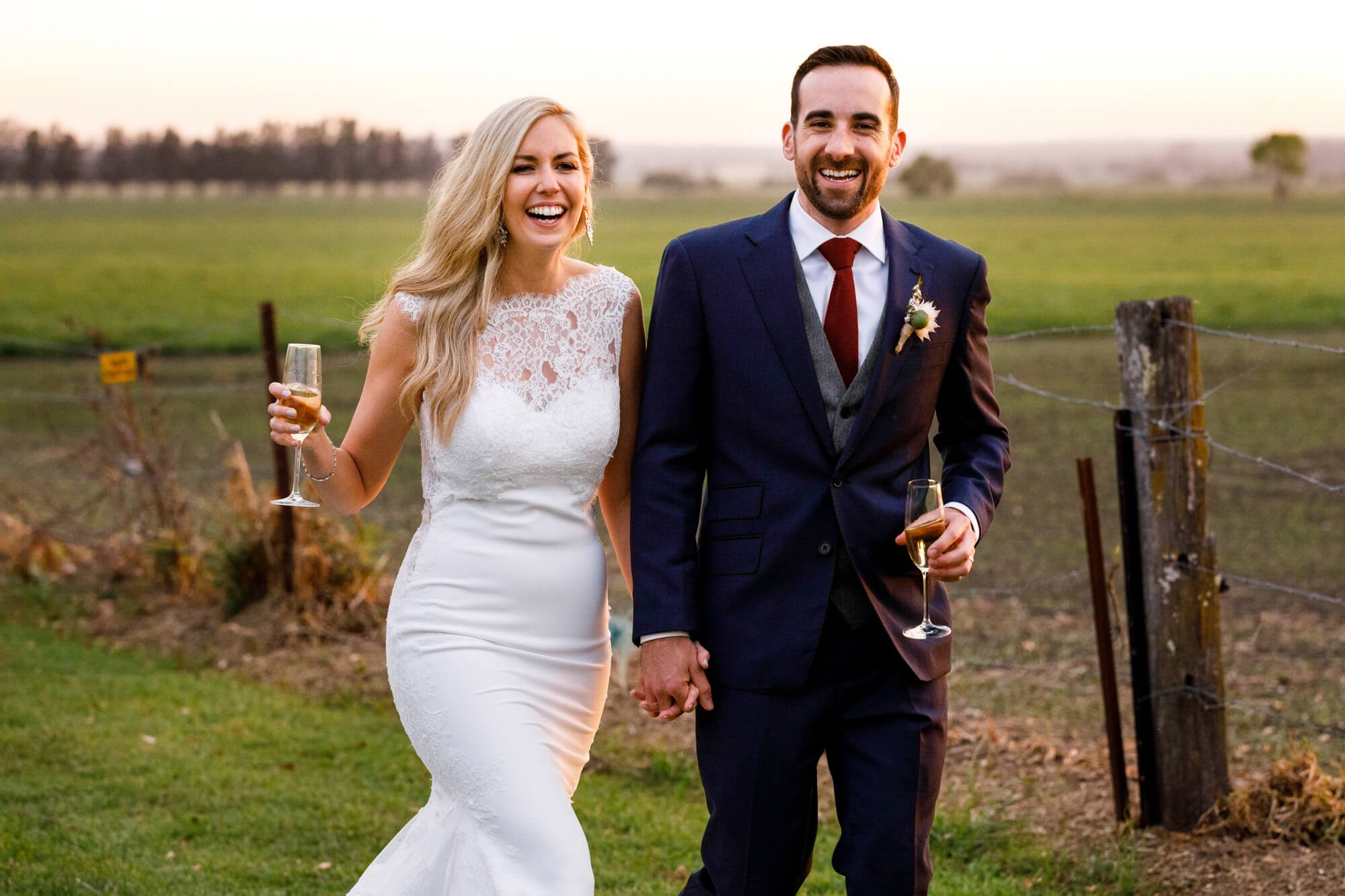 Bride and groom laughing together as they walk hand in hand across open farmland, holding champagne glasses, Merribee House, South Coast, NSW