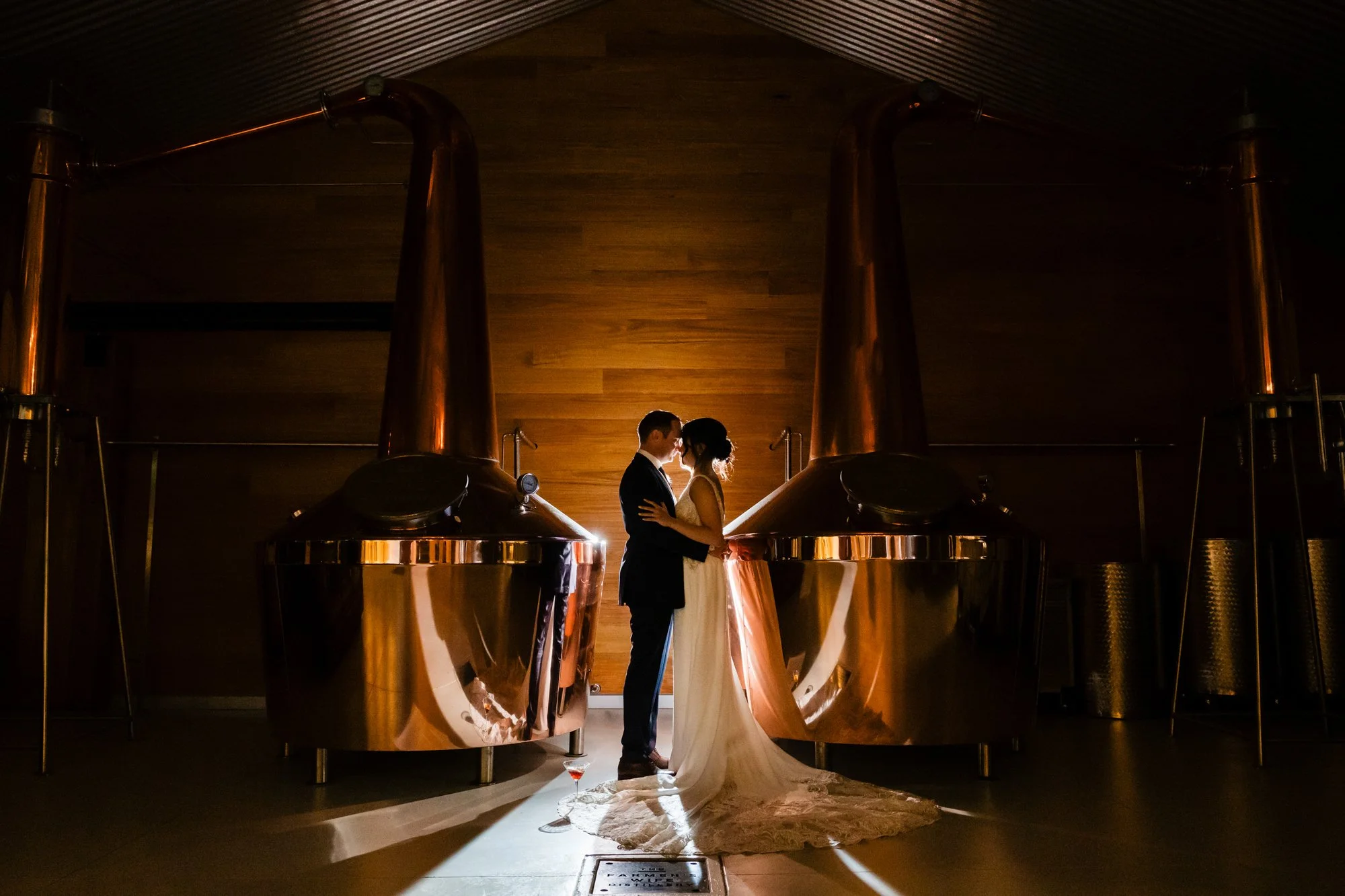 Bride and groom slow dancing together beside towering copper brewing tanks, warm light glowing over the intimate moment, The Farmer's Wife Distillery, Allworth