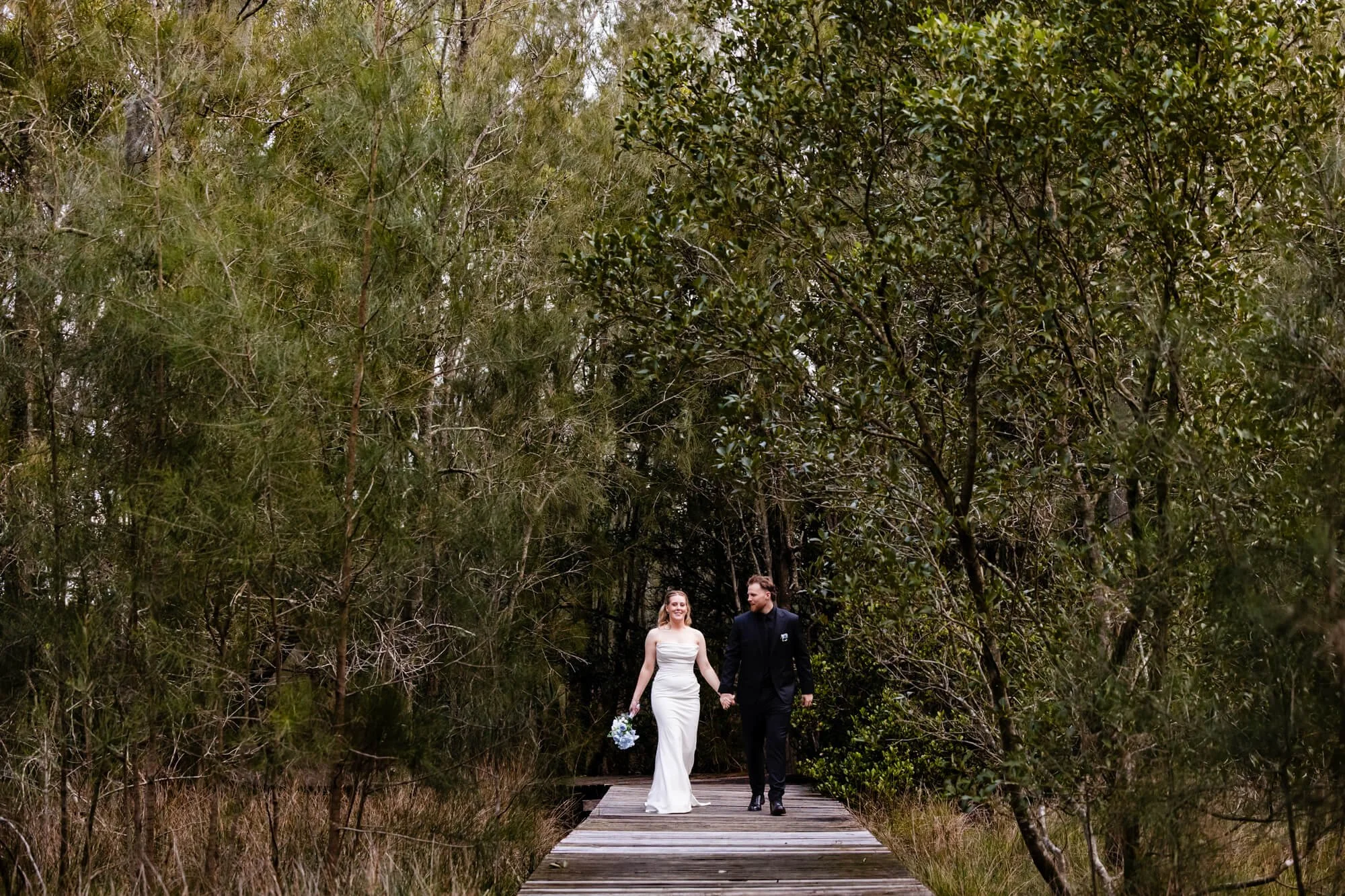 Bride and groom walking hand-in-hand along a wooden boardwalk through lush rainforest at Heaven Gardens, Port Stephens.